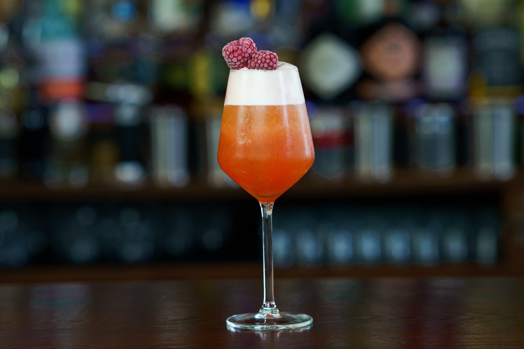A frothy orange cocktail in a wine glass, topped with frozen raspberries, at a bar.