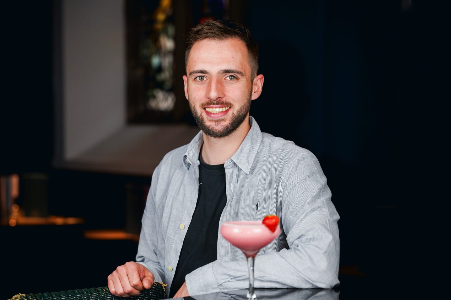 A smiling bartender in a light shirt stands behind a pink strawberry cocktail on the counter.
