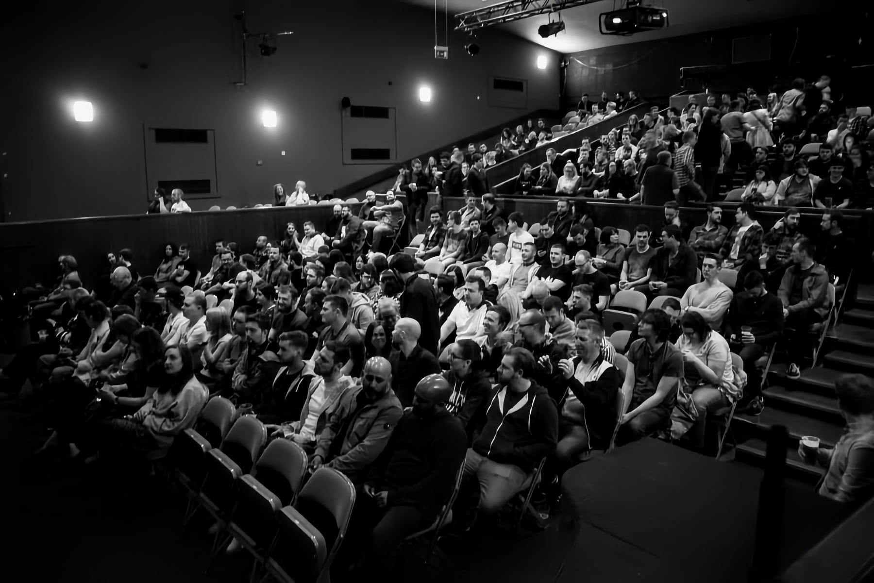 Black and white photo of a busy theatre audience seated and chatting before a performance begins.