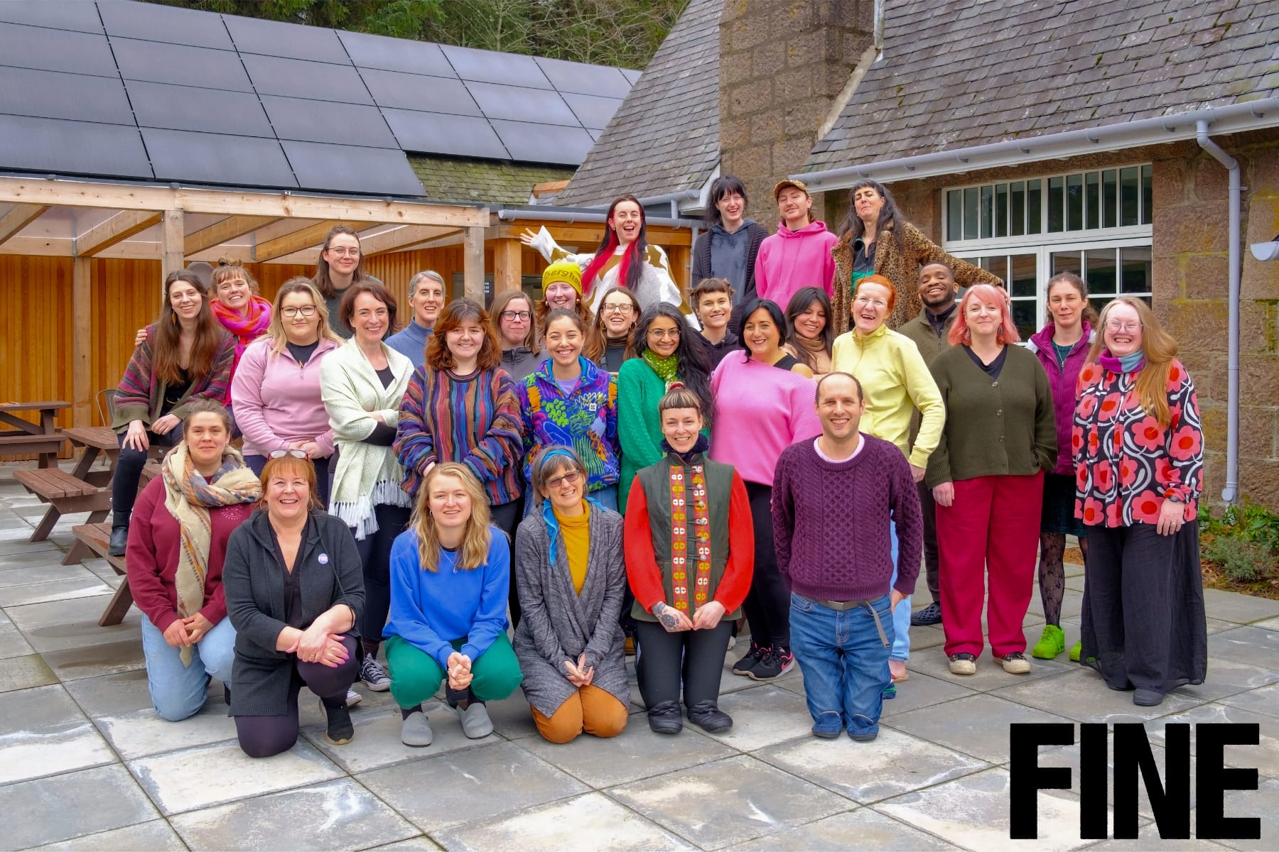 Large group of smiling people in colourful clothes posing outside a stone building.