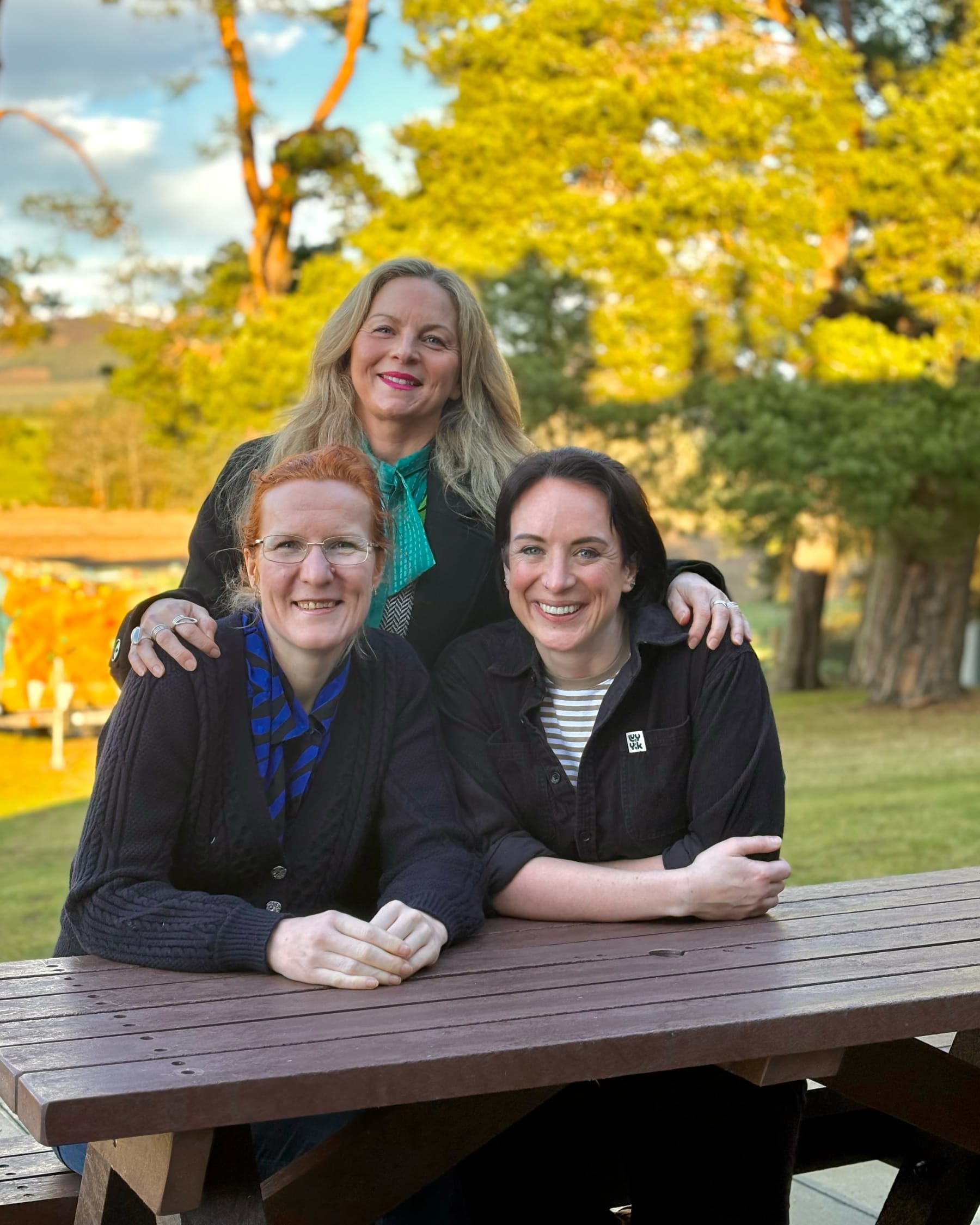 ally Reaper, Birgit Itse and Claire Abbott smiling at a picnic bench with trees behind them.