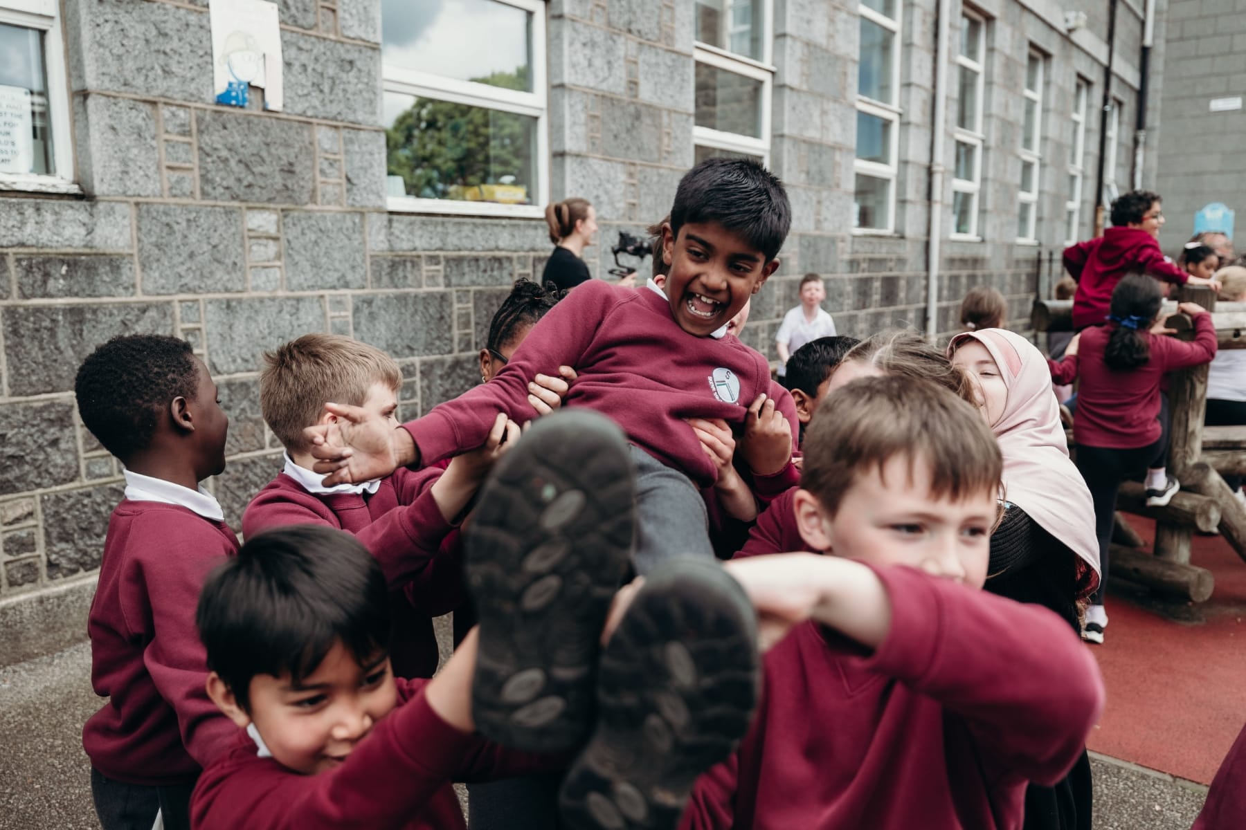 Group of schoolchildren in maroon uniforms lift a joyful classmate in a playground outside school.