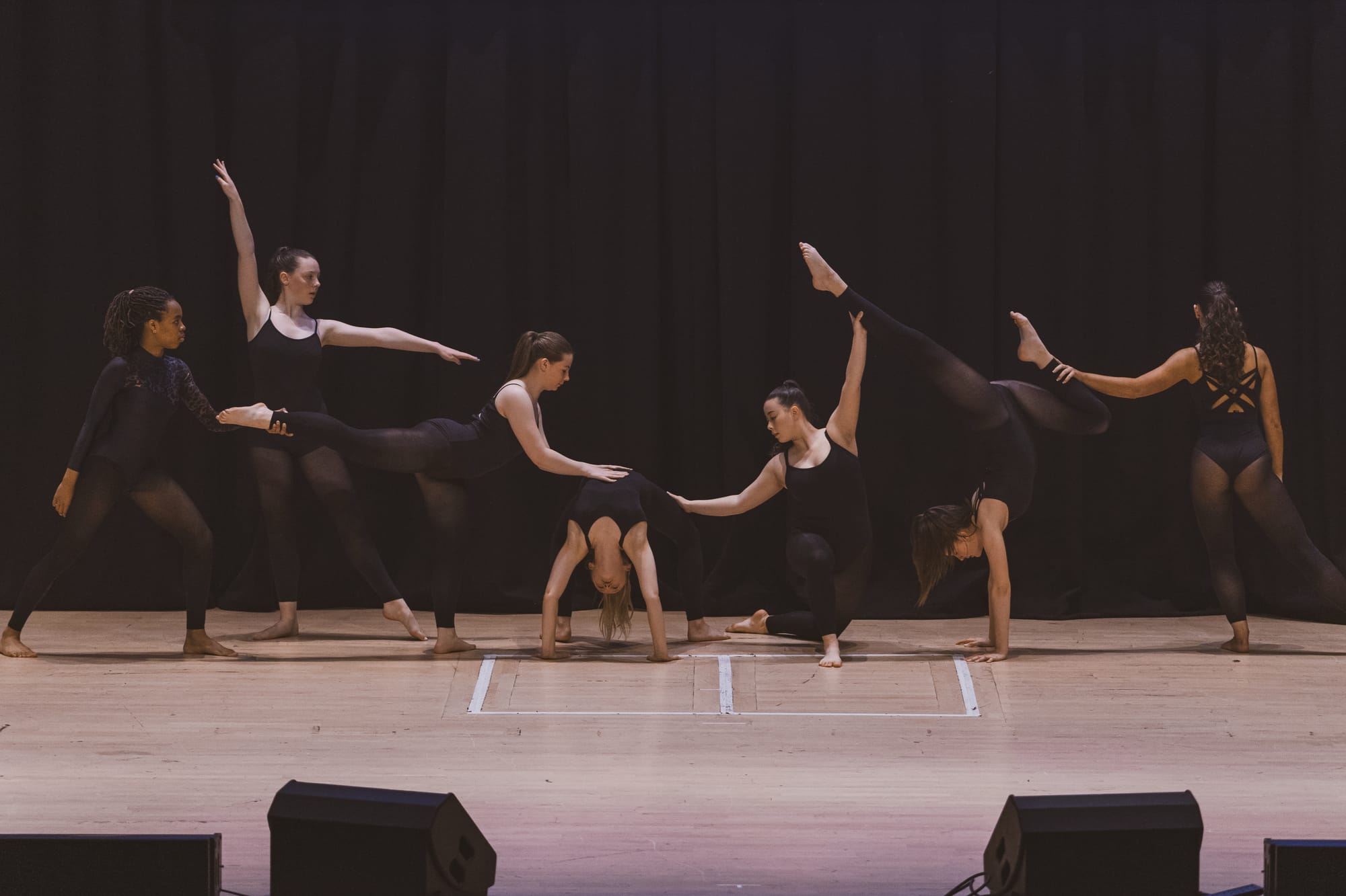 Seven dancers in black costumes perform a contemporary routine on stage with dramatic poses and lifts.