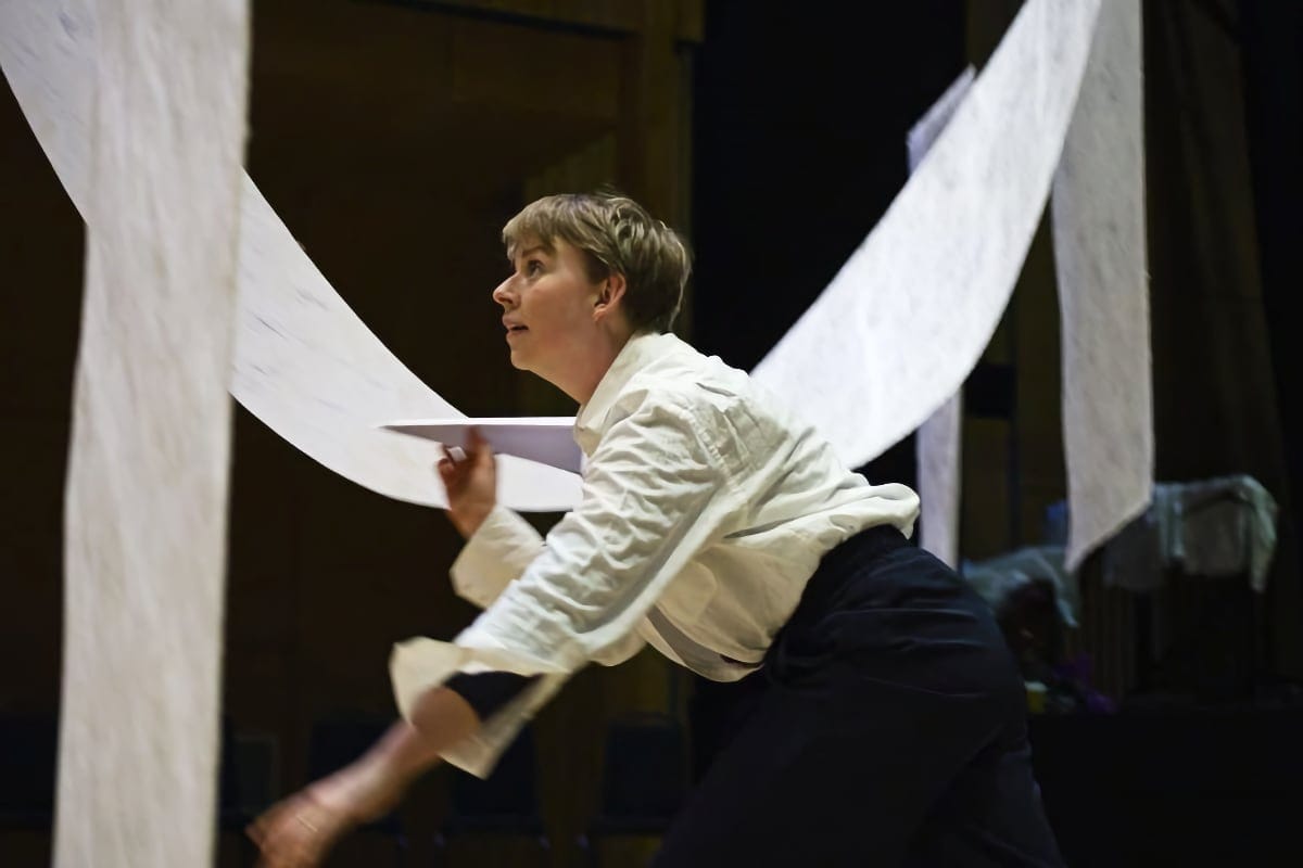 A dancer performs A Journey of Flight, captured mid-motion holding a paper plane beneath floating sheets of paper.