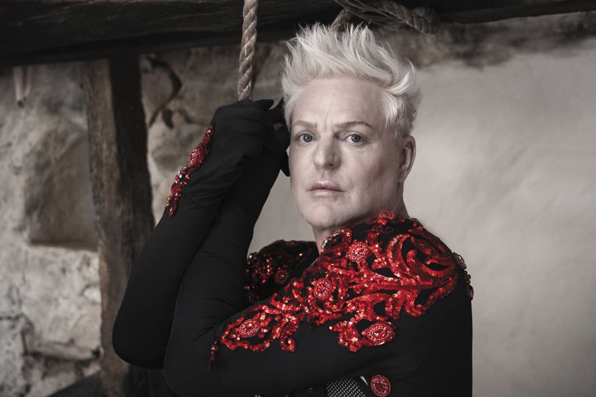 Singer Andy Bell poses in a dramatic setting, wearing a black outfit with ornate red sequinned embroidery and striking platinum hair.
