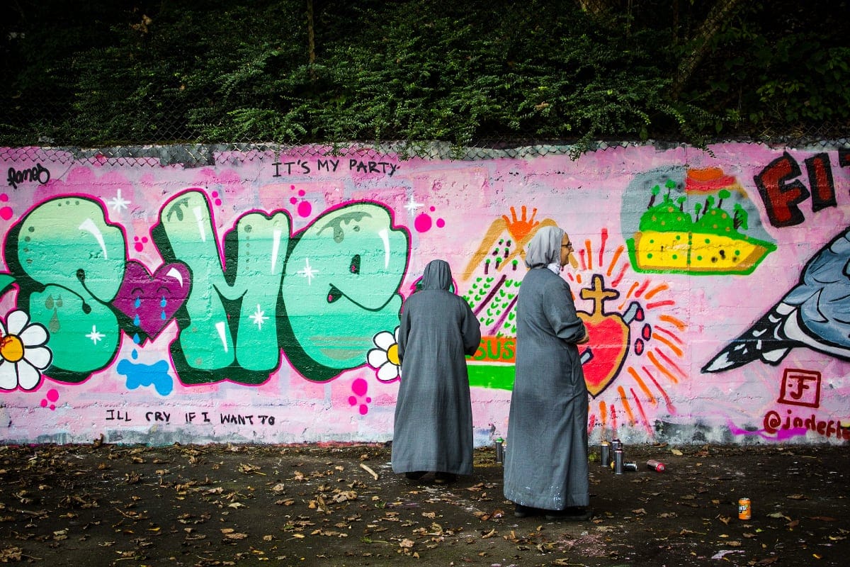 Two nuns in grey habits stand in front of a brightly coloured graffiti wall, which features bold text, flowers, a sacred heart, and cartoon-style art on a pink background.