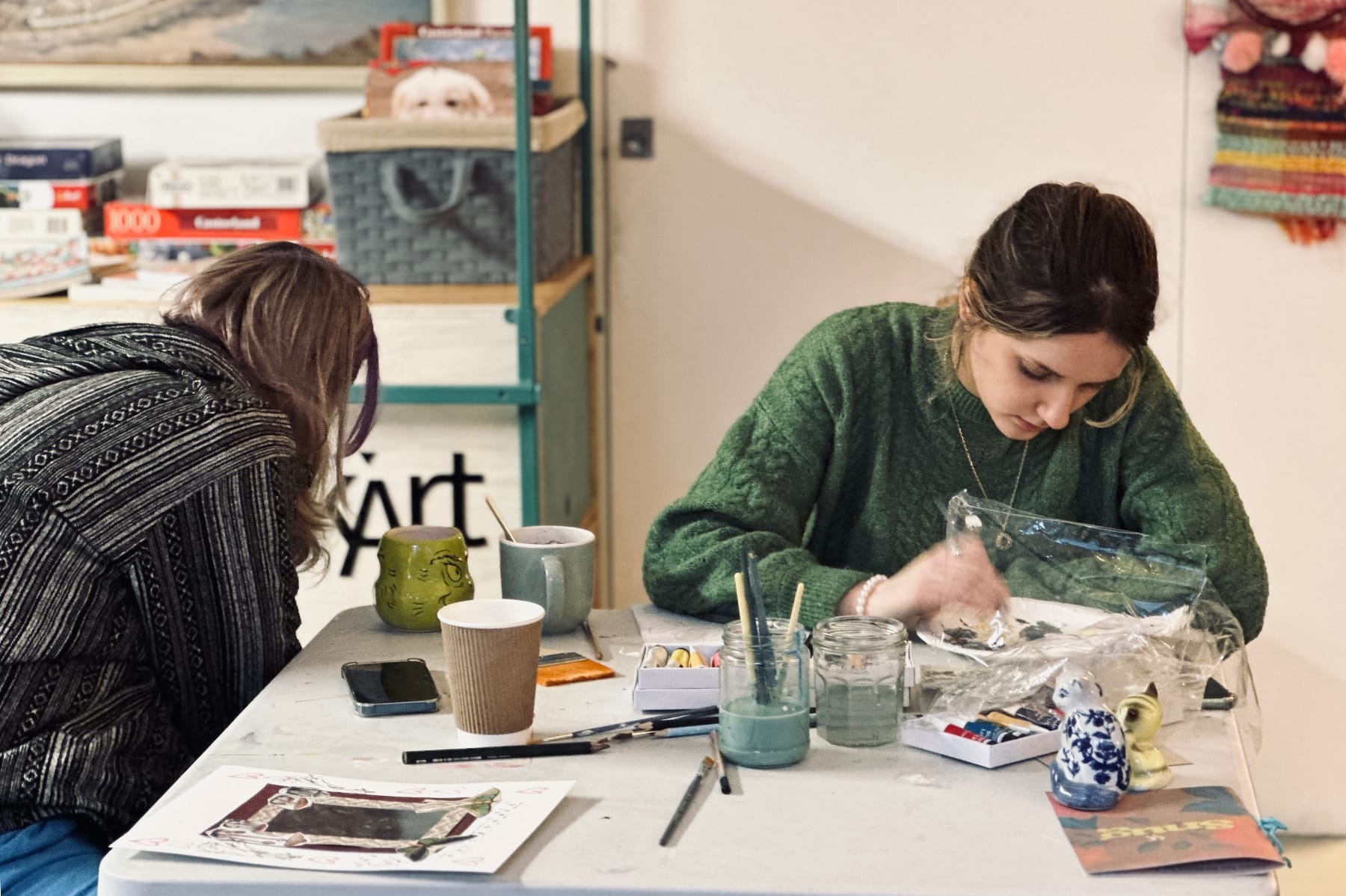 Two women sitting at a table focusing on creative tasks, surrounded by mugs and painting tools.
