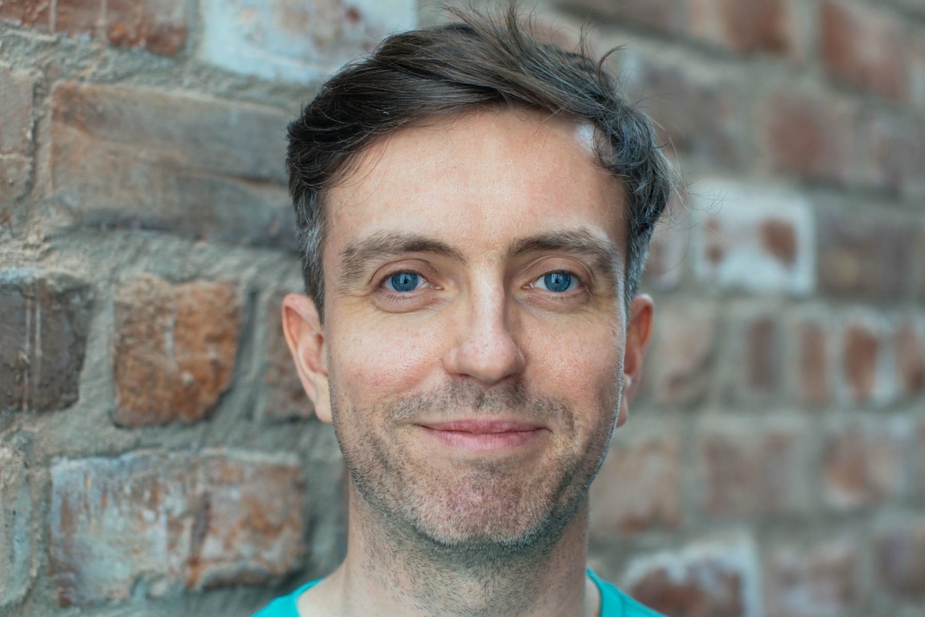 Greg Sinclair standing against a brick wall, smiling, wearing a teal T-shirt