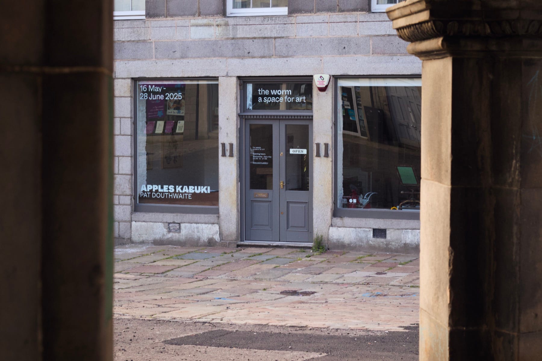 A quiet urban gallery with posters for Apples Kabuki by Pat Douthwaite, 16 May–28 June 2025.