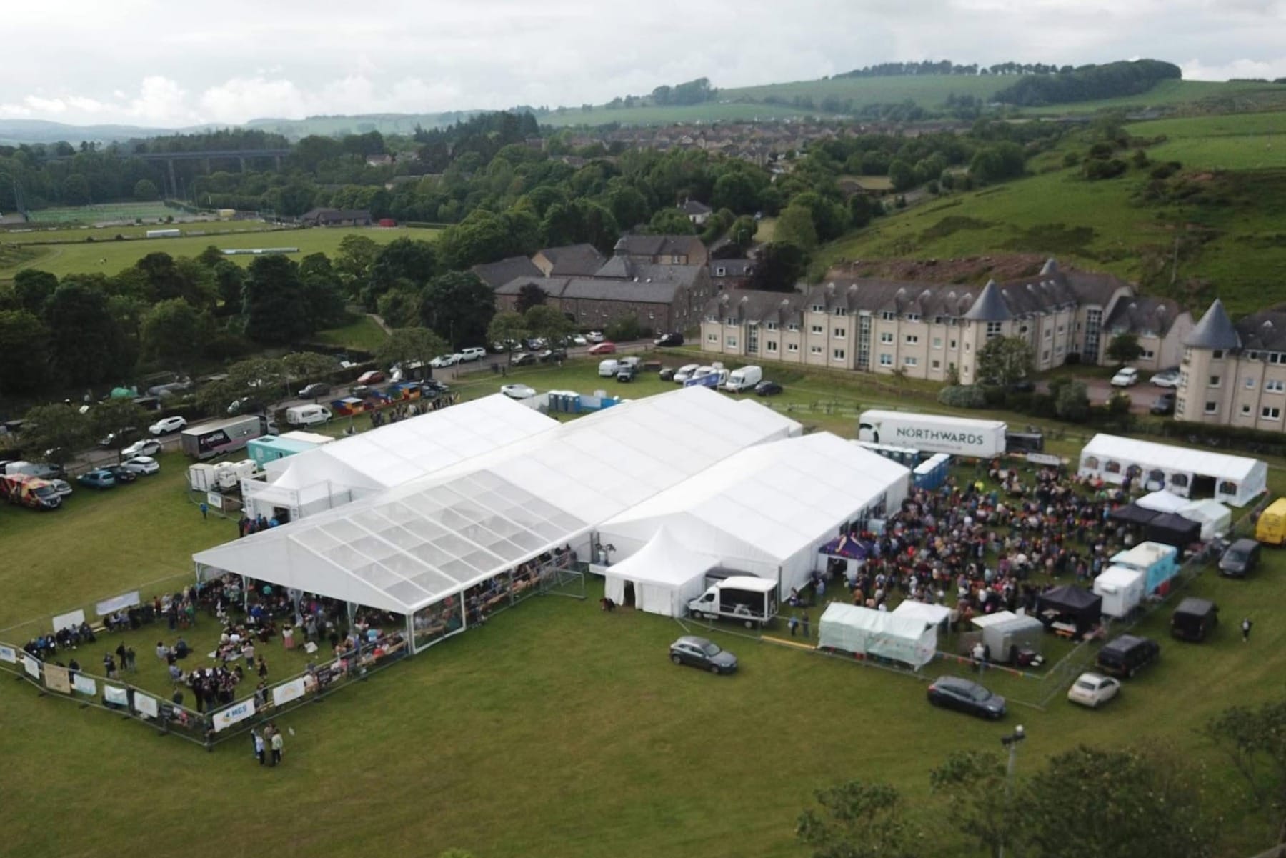 A large white marquee with a clear-roofed section sits on a grassy field surrounded by people, stalls, cars and nearby housing, with green hills and countryside in the background.