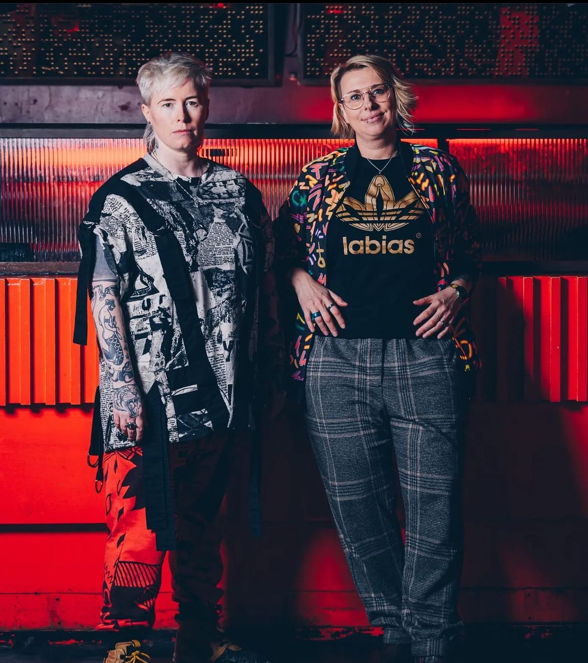 Lezzer Quest stand in front of a red wall wearing bold prints and statement outfits.