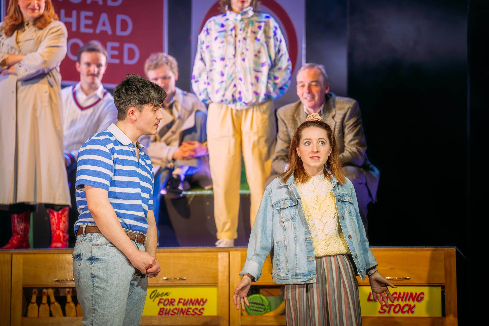 Two young actors in 80s-style outfits speak on stage, with cast members seated behind them.