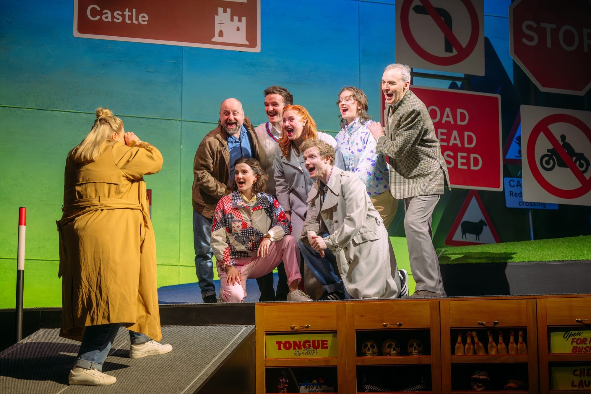 A group of performers pose for a photo in front of playful road sign props on a theatre stage.