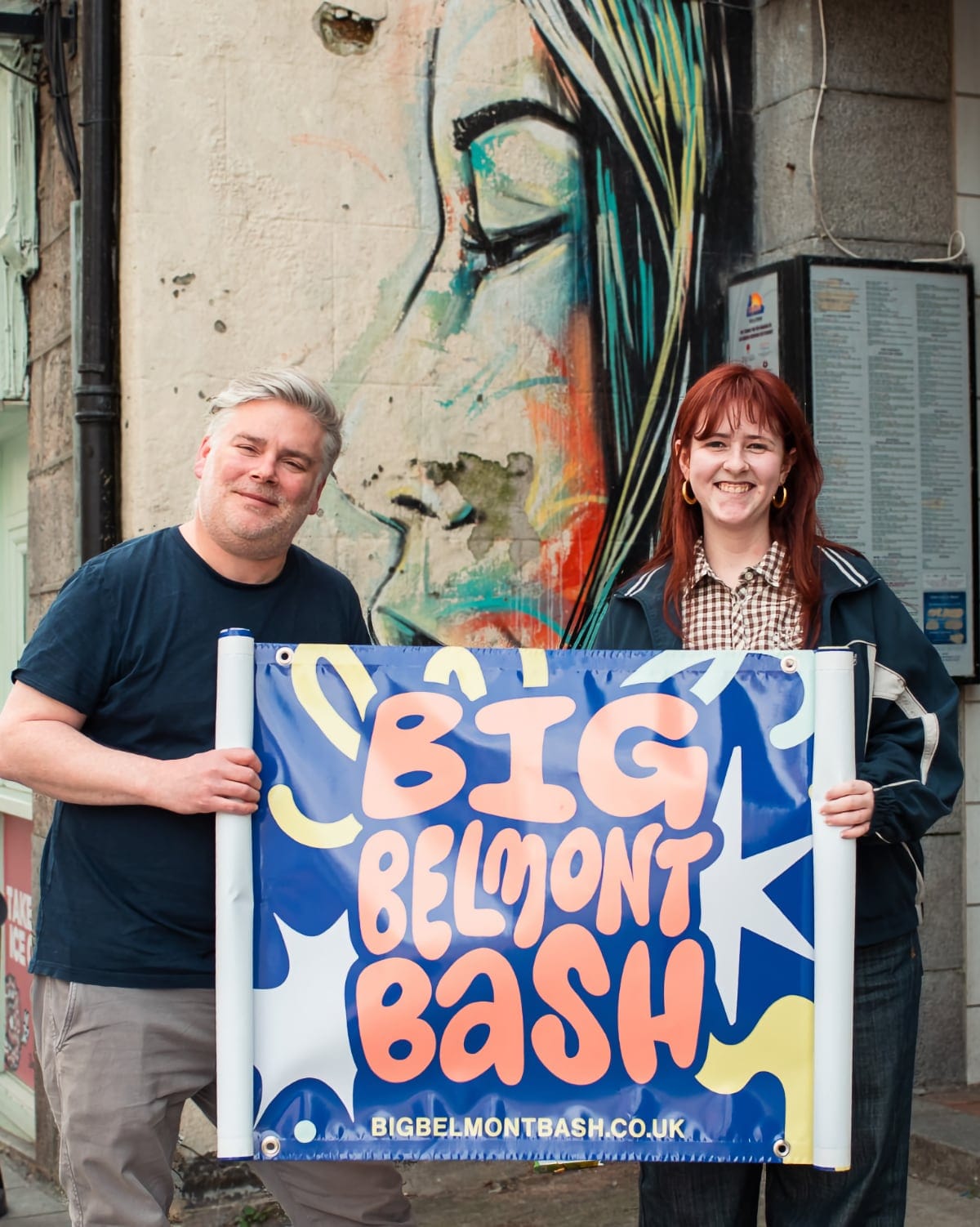 Two people stand smiling outside on Belmont Street holding a banner that reads “Big Belmont Bash.” Behind them is a brightly painted mural of a woman’s face with closed eyes and flowing hair.