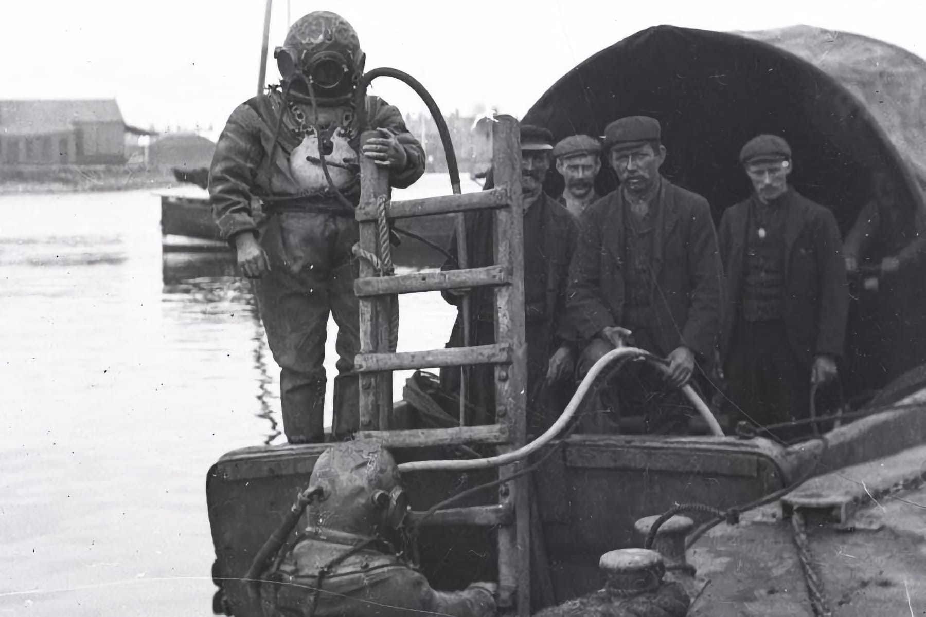 Early 20th-century deep-sea divers with heavy gear and hoses, preparing for or emerging from a dive, surrounded by workers on a dock.