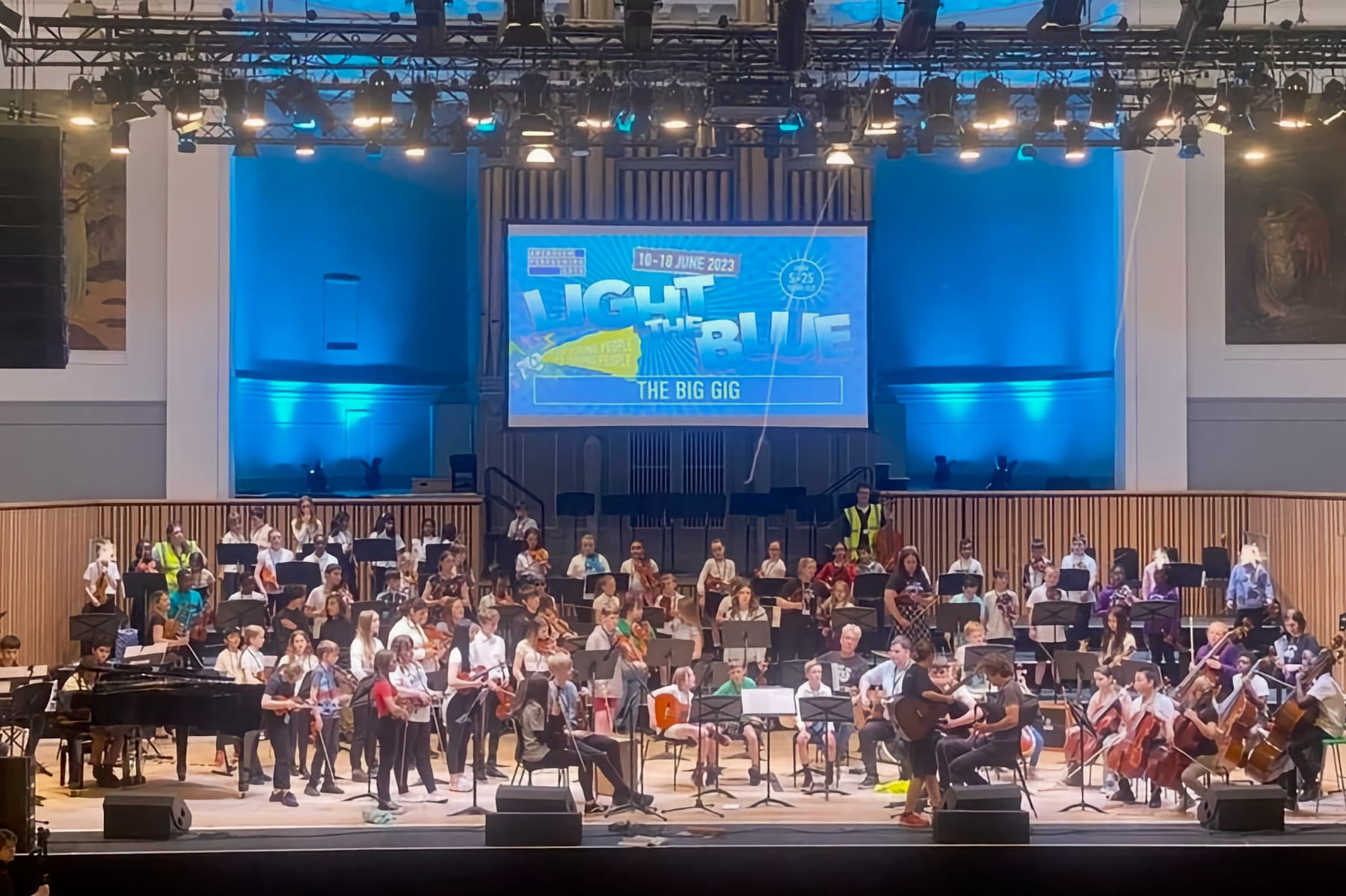 Light the Blue’s “The Big Gig” in Aberdeen’s Music Hall, where young musicians and singers are preparing for a performance. The hall is bathed in blue lighting and features a prominent screen with the event’s name