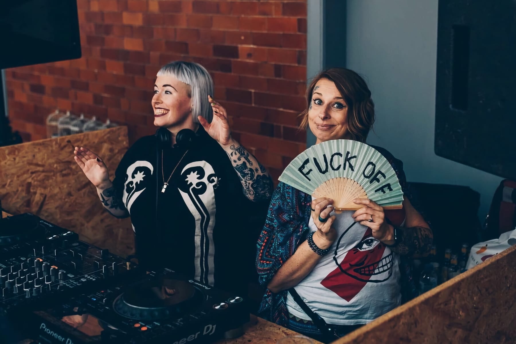 Lezzer Quest members stand smiling, one holding a “Fuck Off” fan while DJ equipment is nearby