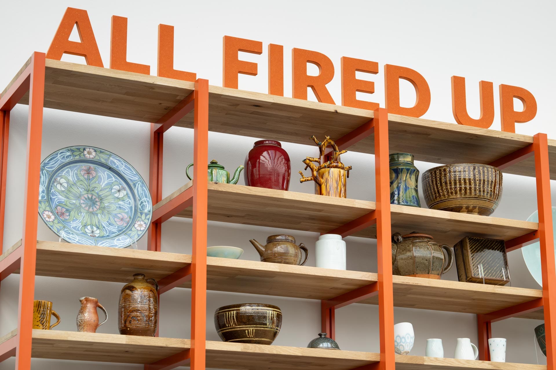 Wooden shelves with orange frame hold colourful ceramic vessels under bold orange letters reading ALL FIRED UP.