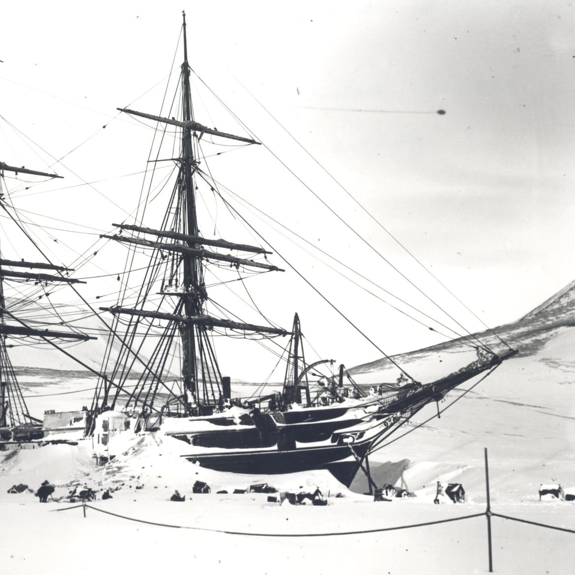 Black and white photo of a tall ship with three masts, frozen in thick Antarctic snow with sled dogs nearby.