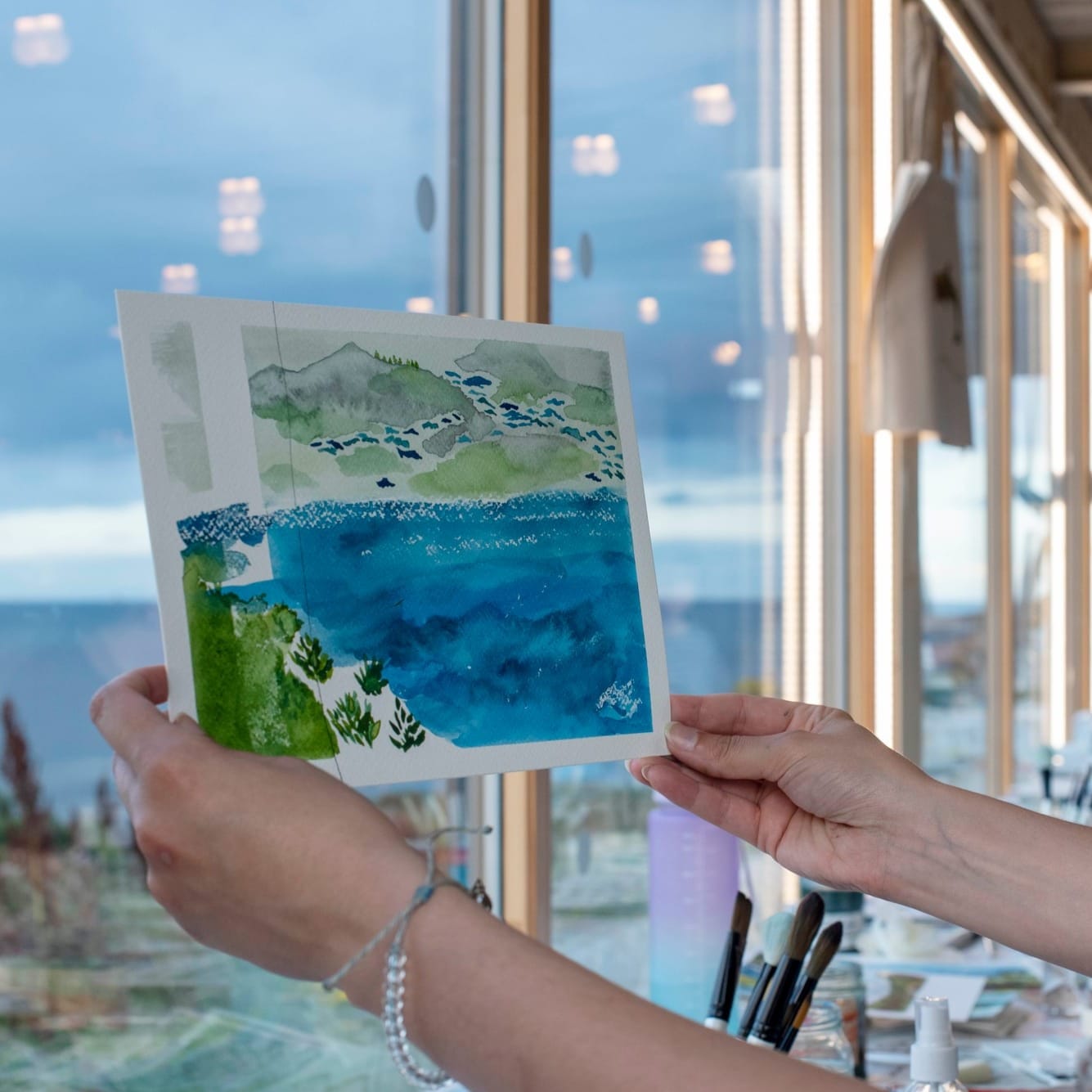 Hands hold a watercolour painting of sea and hills in front of large windows overlooking the coastline.