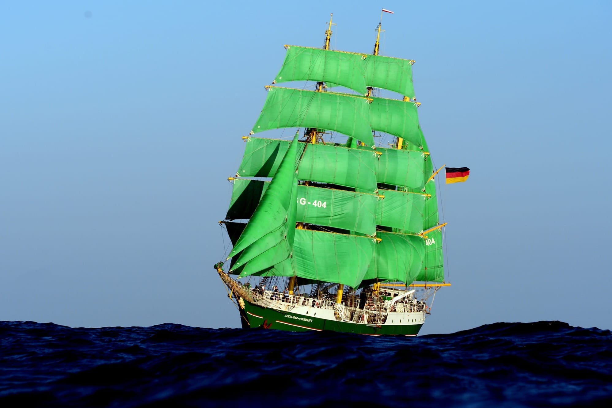 A tall ship with bright green sails, the Alexander von Humboldt, sails on open water under a clear sky, flying the German flag.