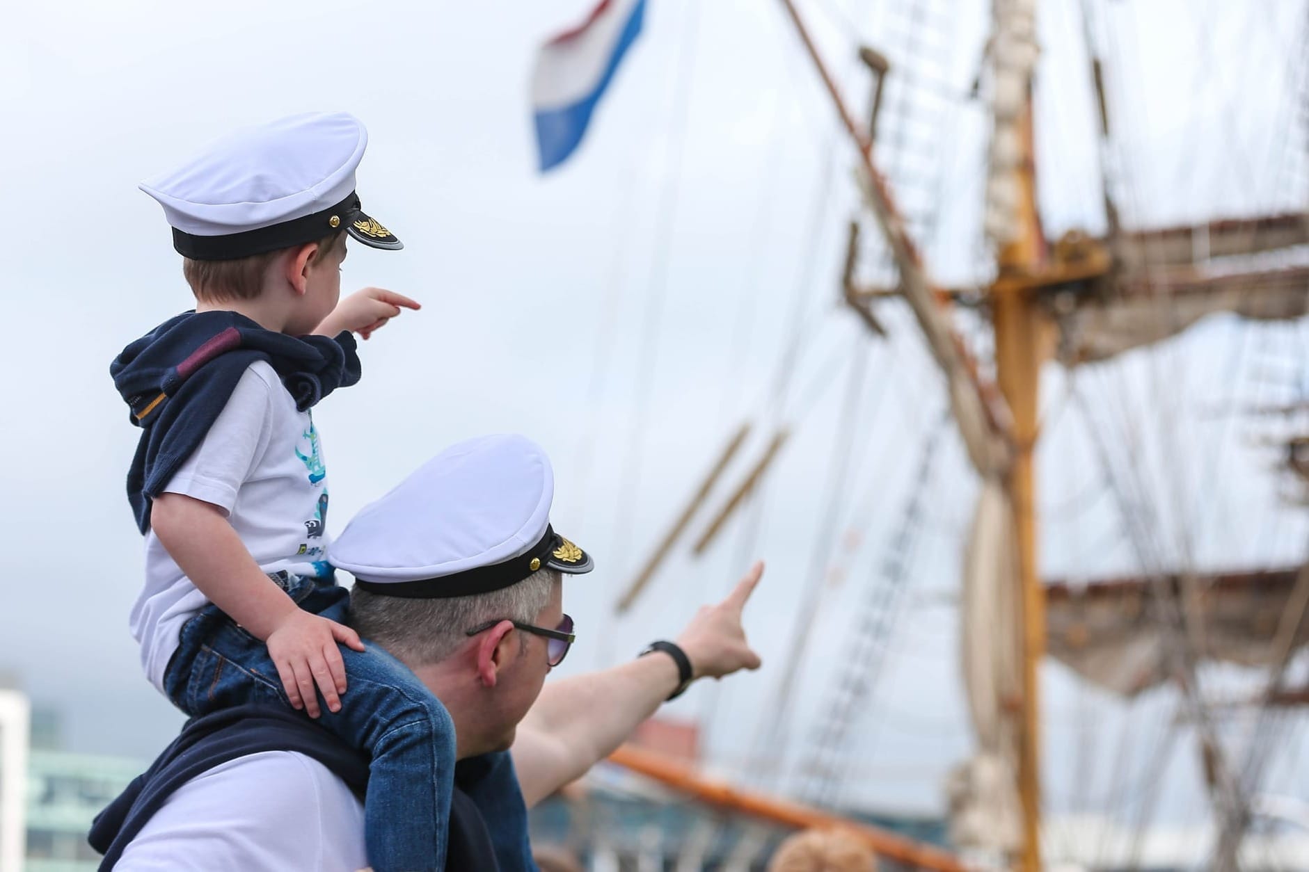 A young child in a captain's hat sits on an adult's shoulders, both pointing excitedly toward a tall ship in the background.