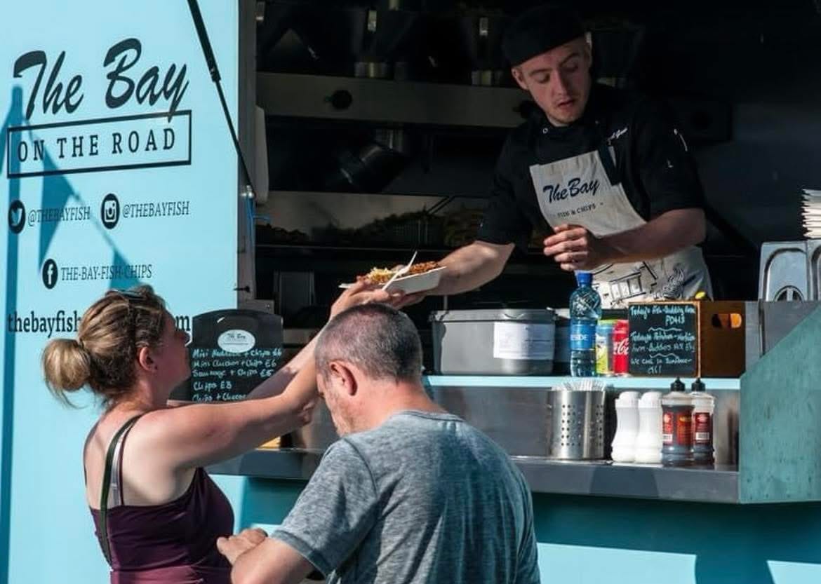 Food truck worker serves a customer from The Bay on the Road van under bright sunlight.