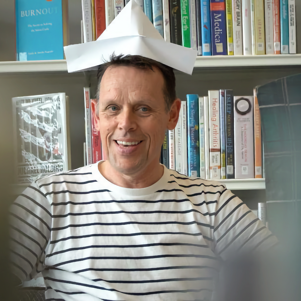A smiling man in a striped shirt wears a paper boat hat, sitting in front of bookshelves in a library.