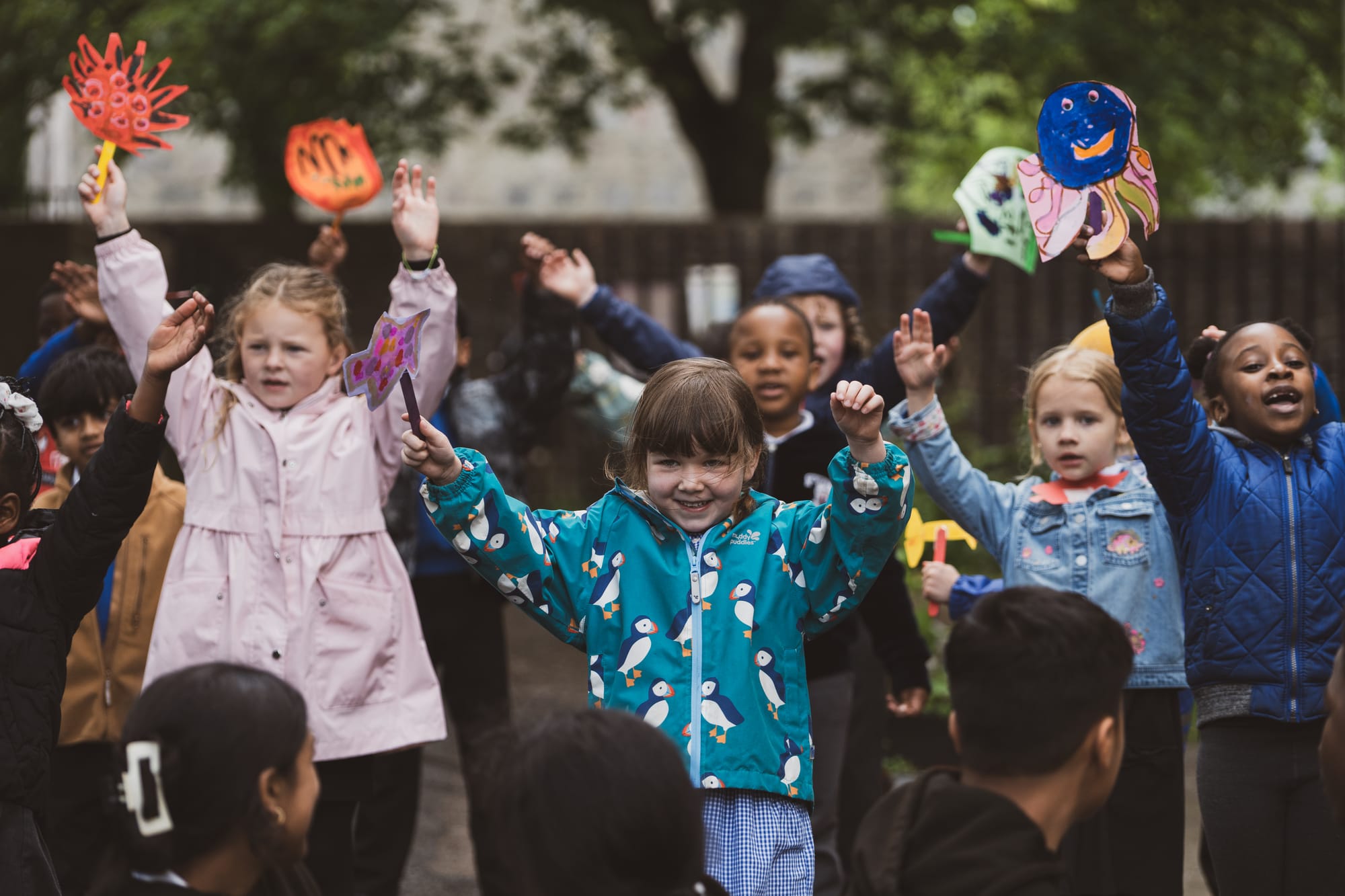 oung children outside raising their arms, holding colourful handmade puppets and artwork.