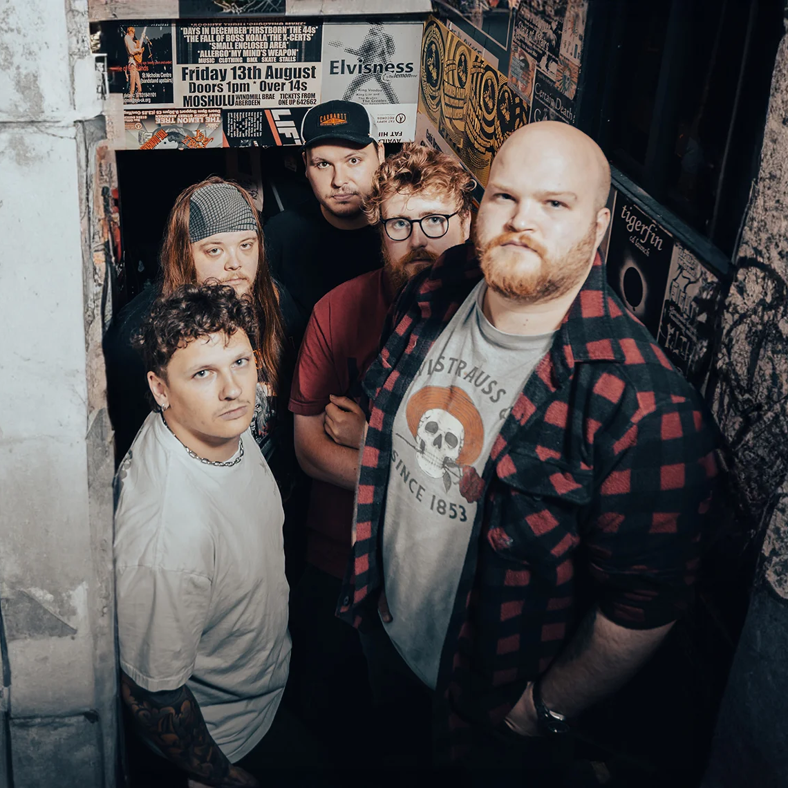 Five members of Atomic Mother stand in a narrow stairwell covered in gig posters and graffiti.