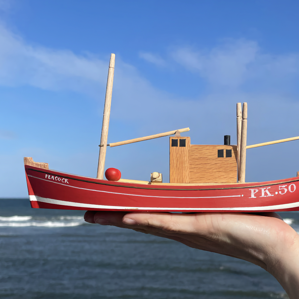 A hand holds a small red wooden model fishing boat marked “Peacock” and “PK.50” in front of the sea.