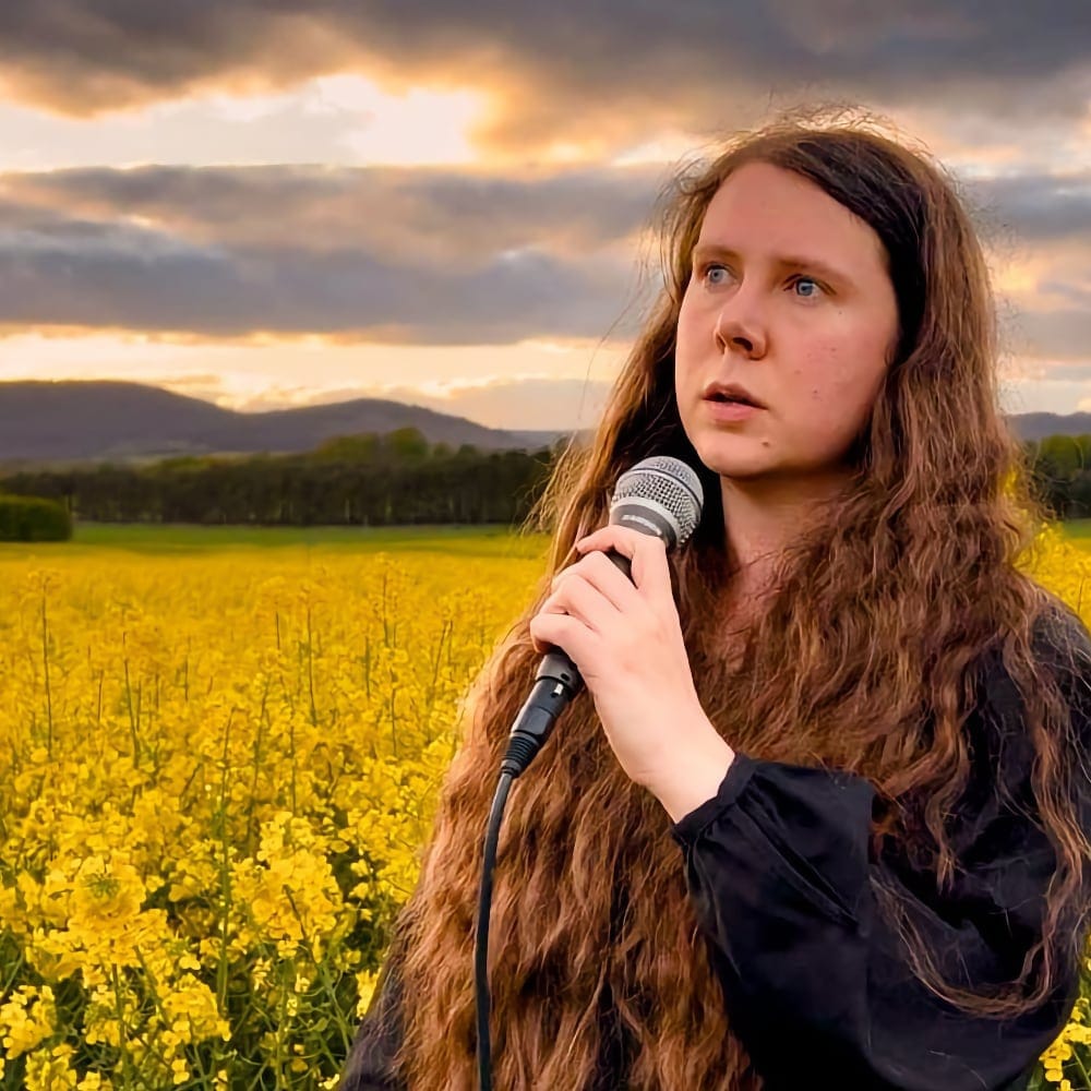 Lucy Beth stands in a yellow field at sunset, holding a microphone and wearing a black top with long hair loose.