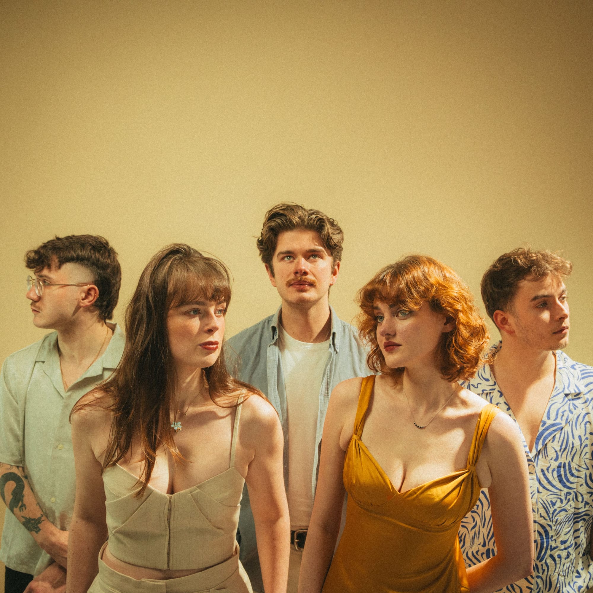 Five band members stand close together in soft lighting against a plain beige background, each looking in a different direction.
