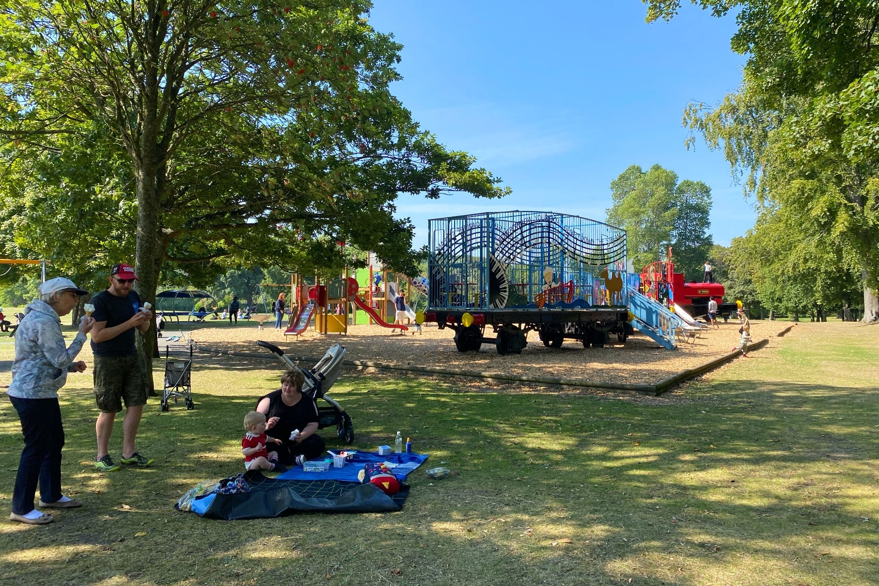 A family picnic on grass near a large playground while others play and walk nearby.