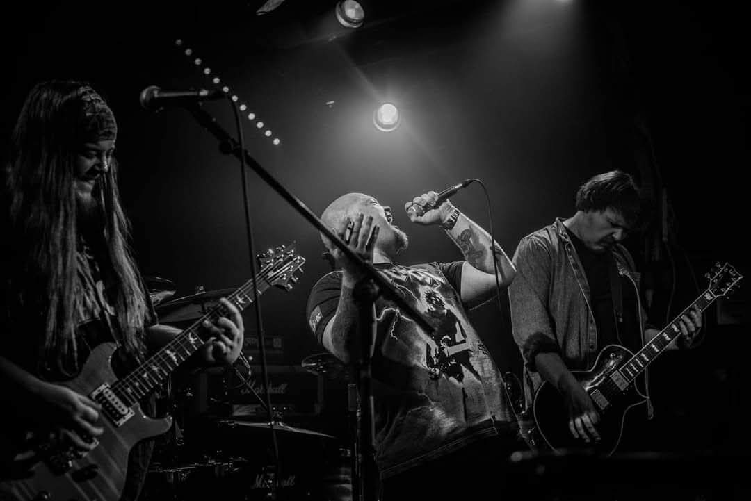 Atomic Mother perform on stage, singer shouts into mic as guitarists play under stage lights.