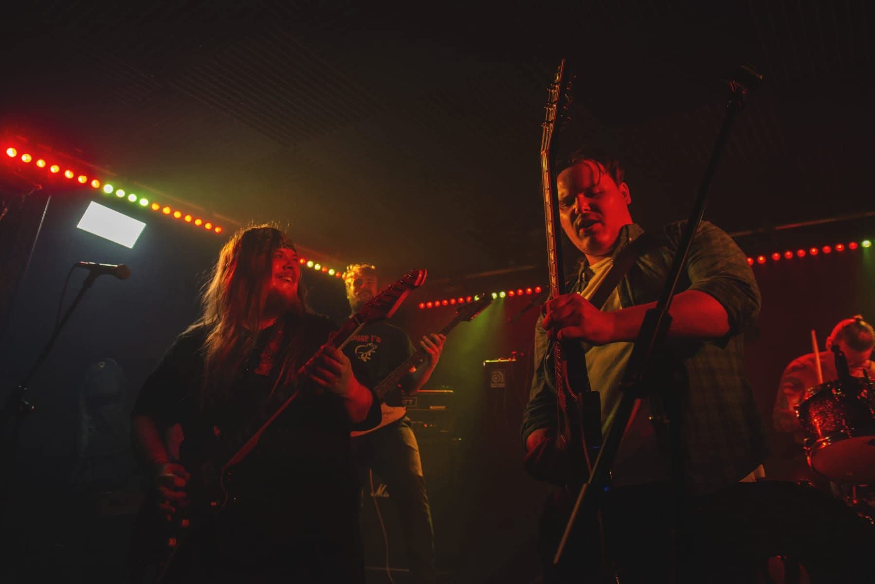 Atomic Mother playing live with red lights, guitarists exchanging looks and drummer in background.