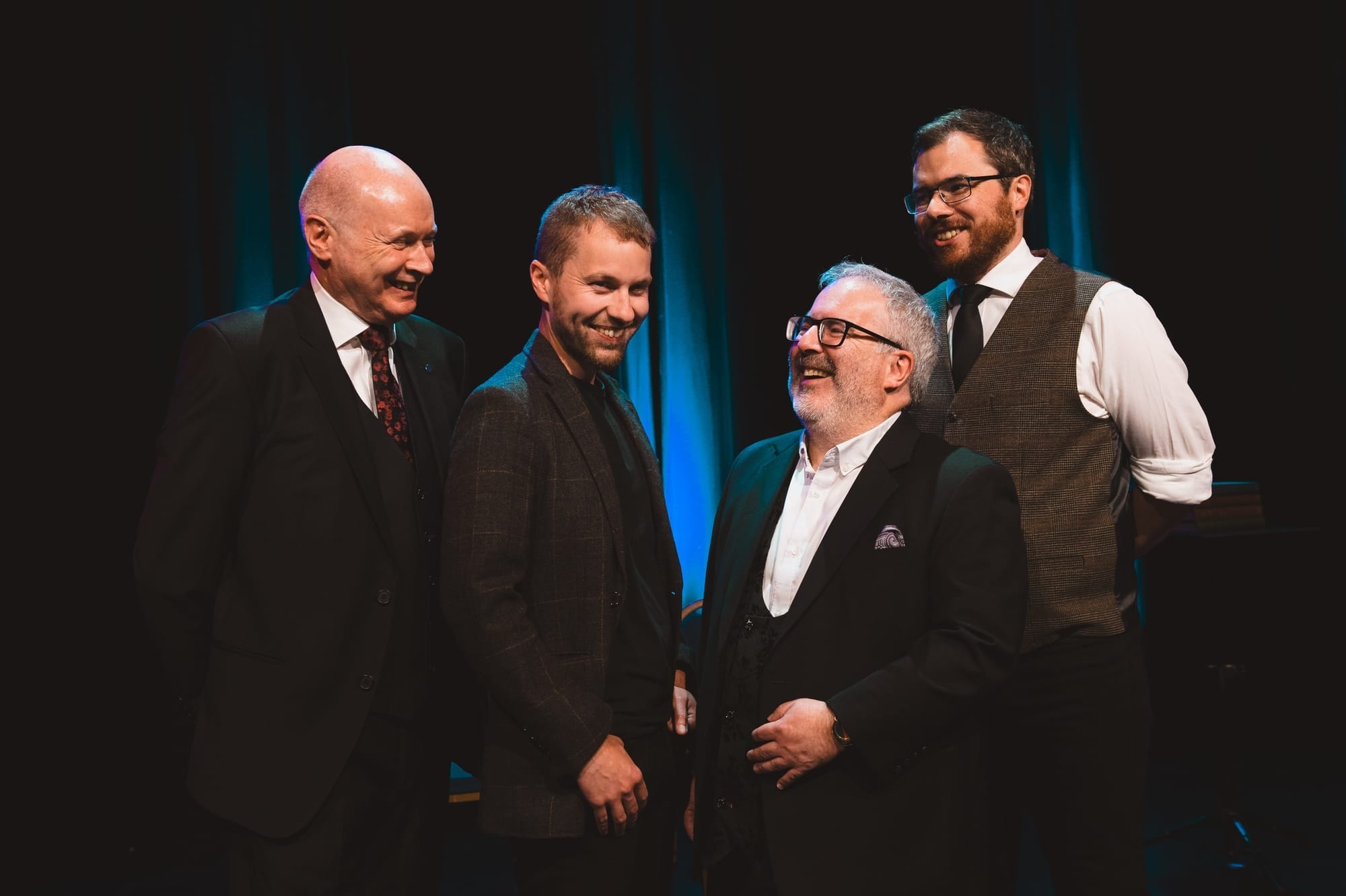 Four smiling magicians in suits on stage, performing at Aberdeen Arts Centre