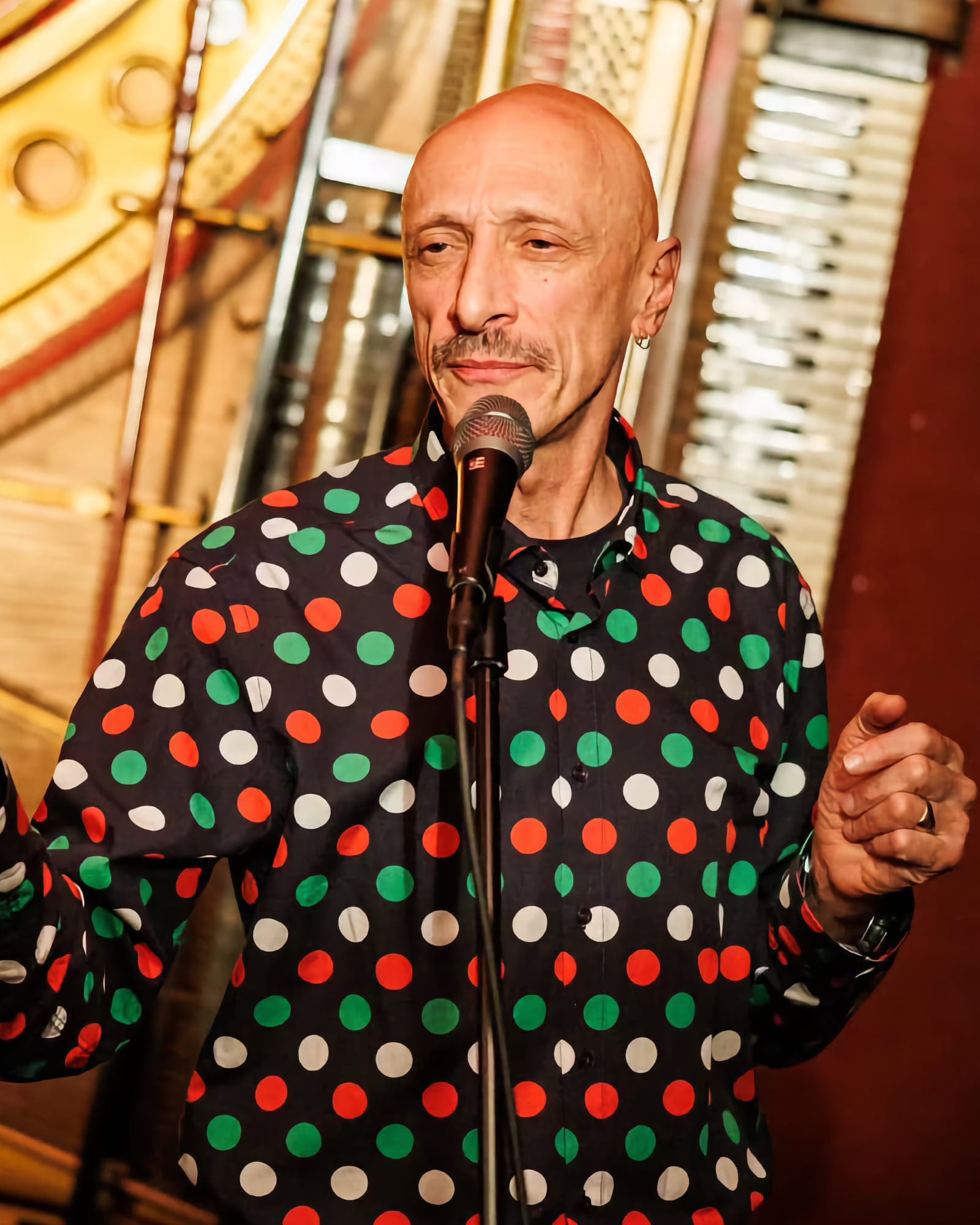 Turan Ali stands at a mic wearing a colourful polka dot shirt, speaking with a thoughtful expression.