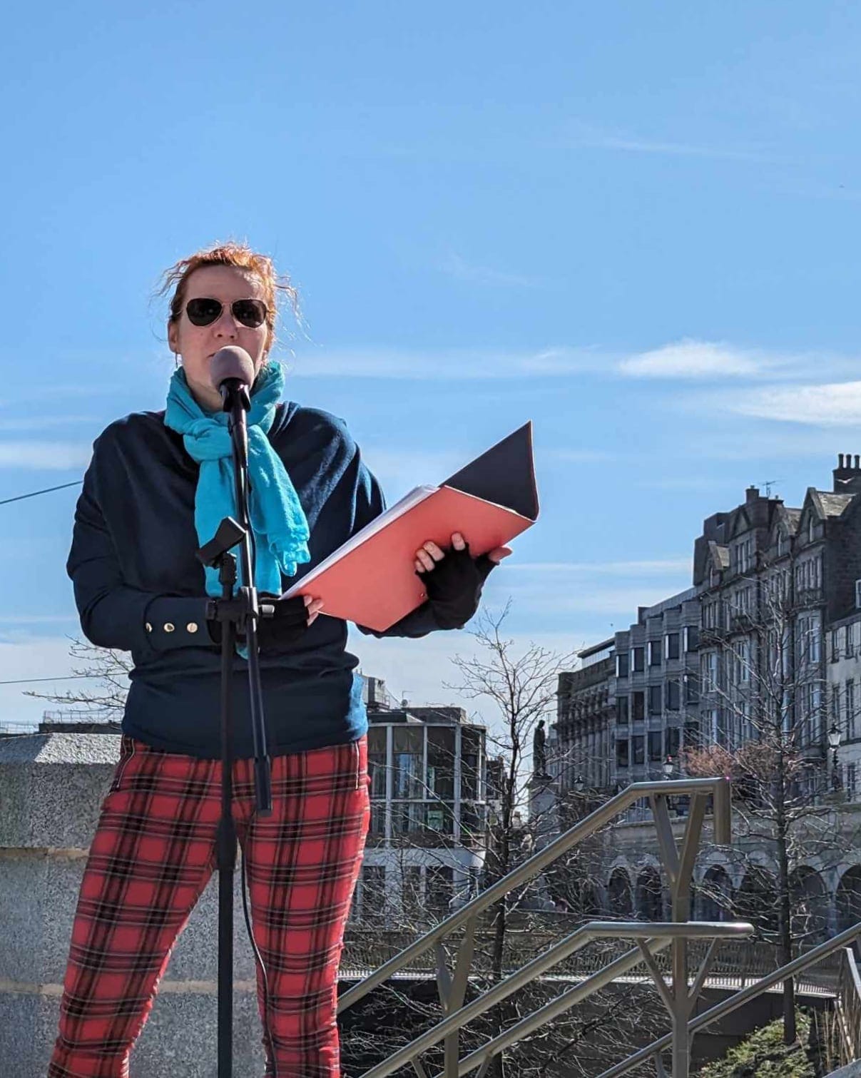 Birgit Itse reads from a folder at a mic, wearing tartan trousers and a blue scarf.