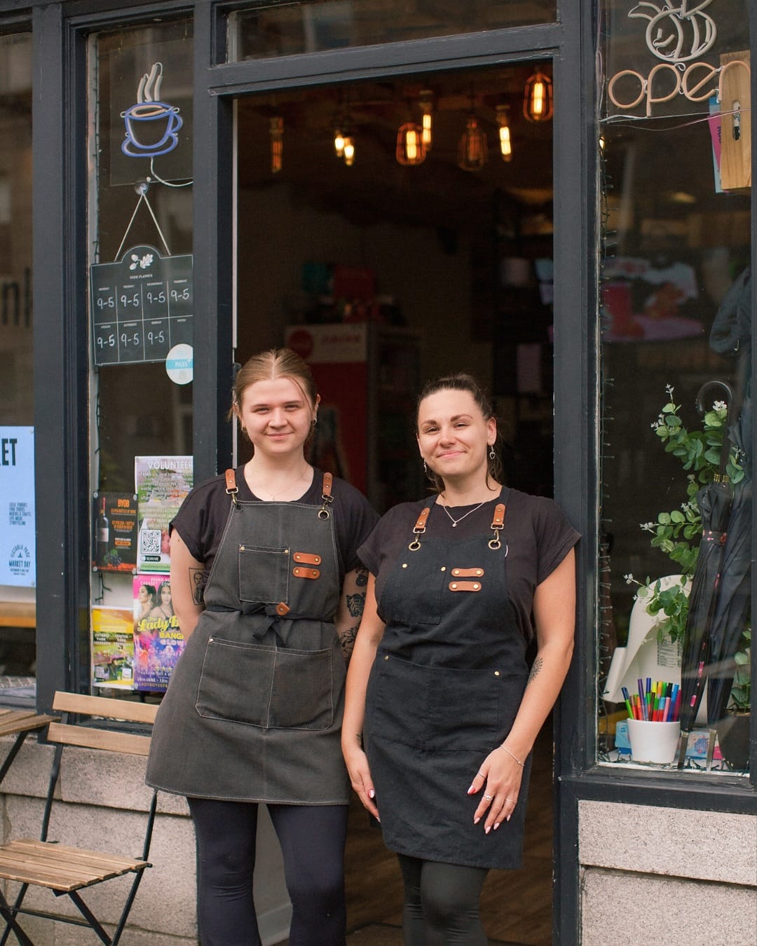 Two team members stand smiling outside Bagel Society, wearing dark aprons in front of the café.