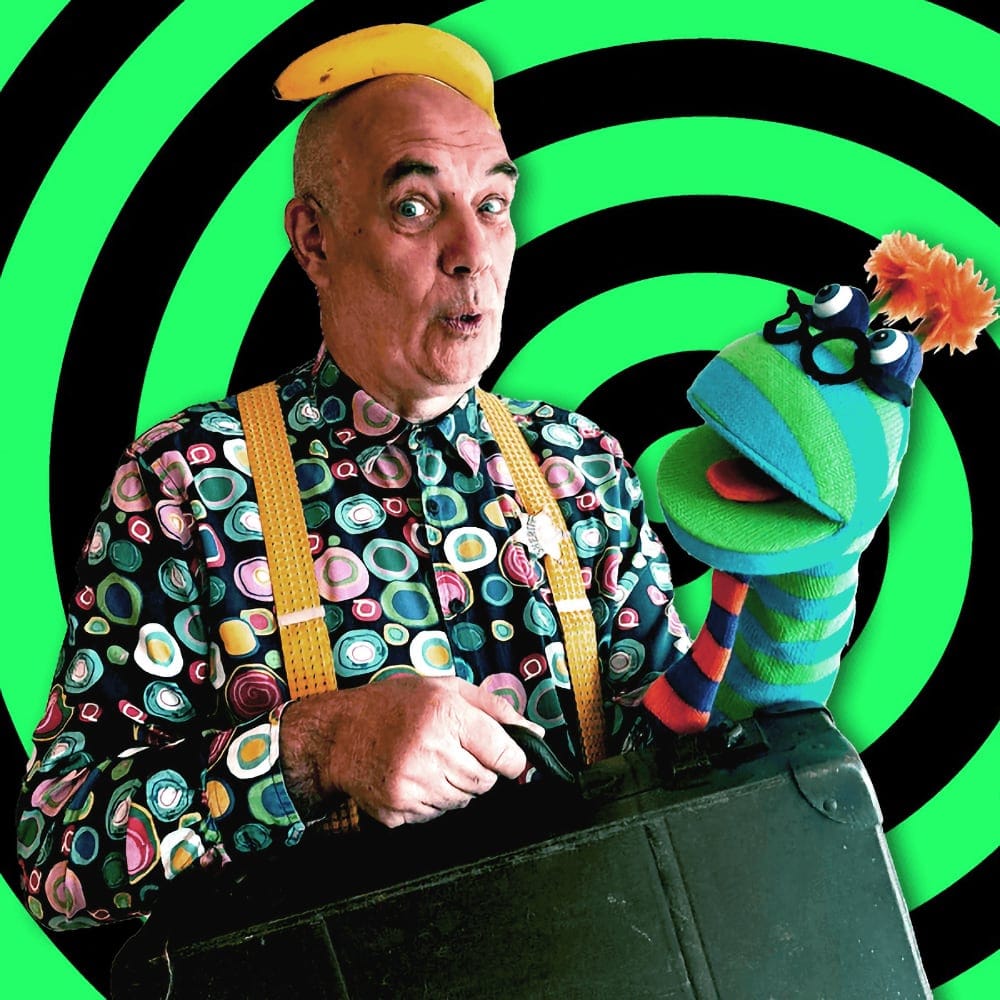Sid Ozalid in a colourful shirt and yellow braces, with a banana on his head and a sock puppet.