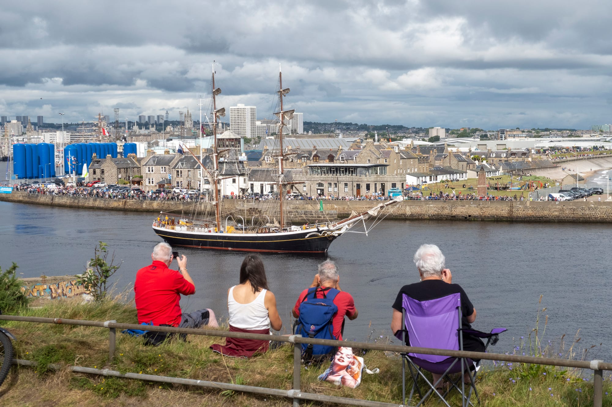 Four people sit on a grassy hill in Torry, watching a tall ship pass through Aberdeen harbour, with crowds gathered along the waterfront in Fittie.