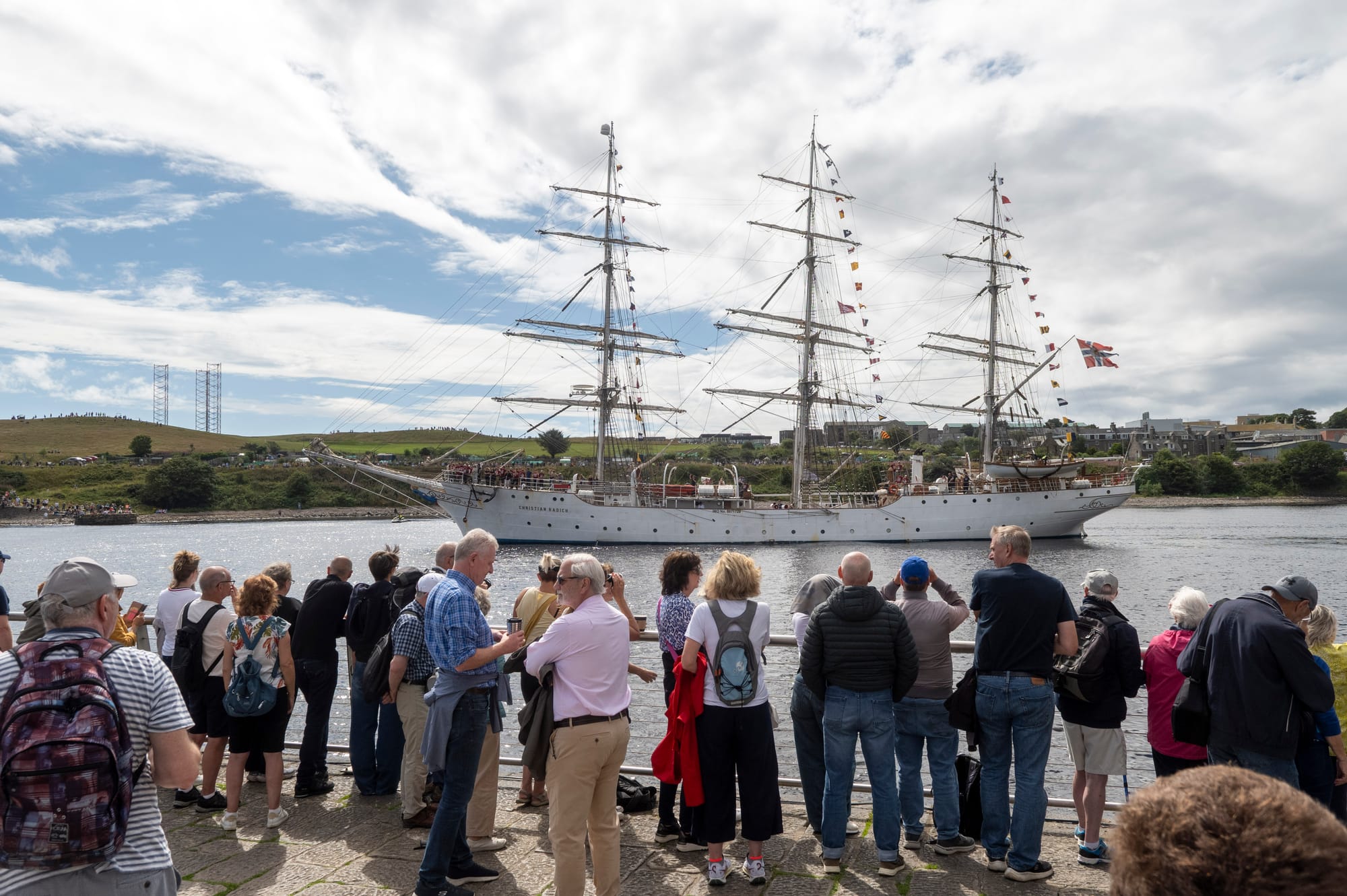 A crowd stands shoulder to shoulder along the waterfront, watching the tall ship Christian Radich sail past on a bright day.