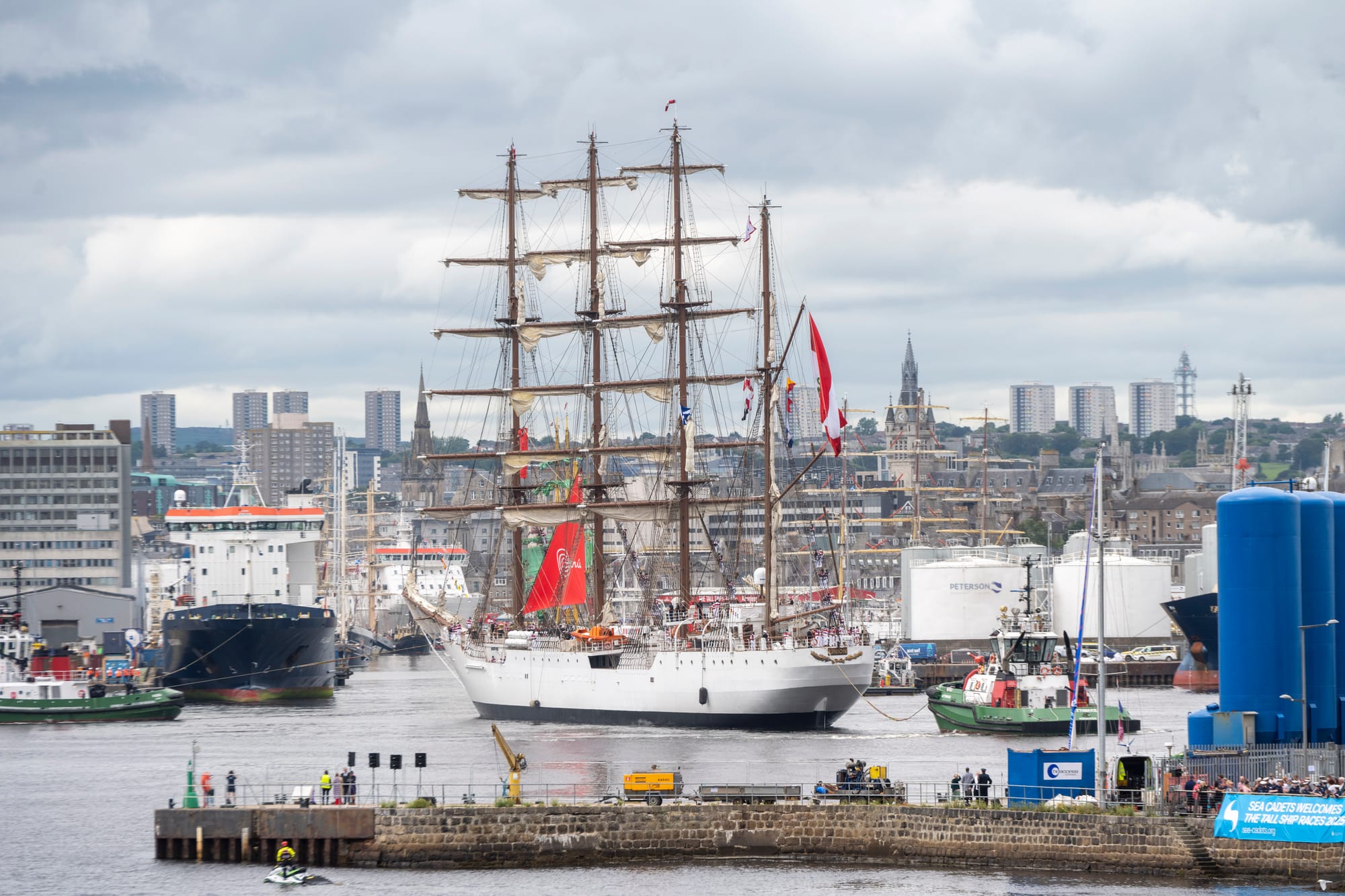 A very large tall ship exits Aberdeen Harbour, surrounded by other vessels, with the city skyline and spires in the background.