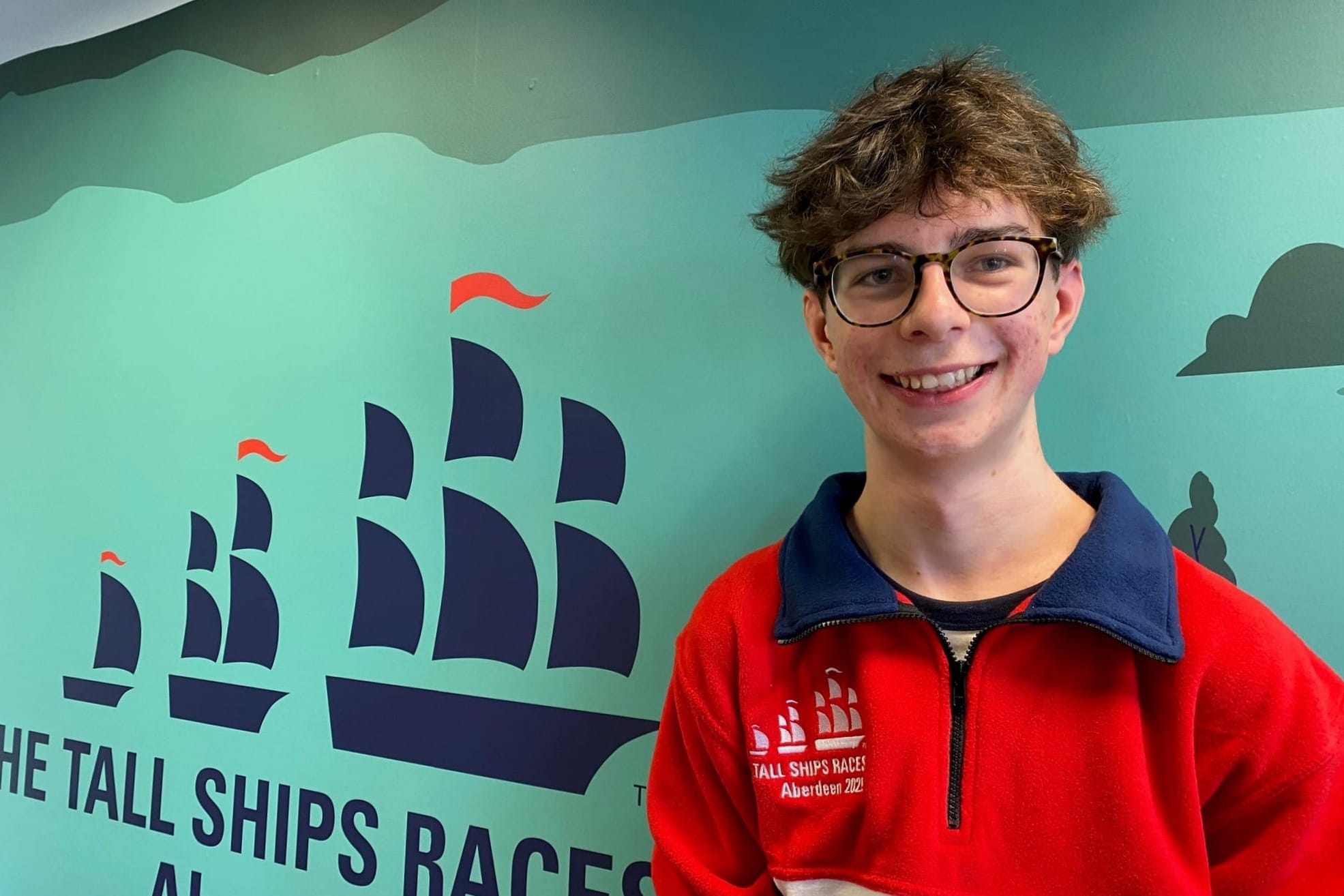 Archie Sweetnam stands in front of the Tall Ships Races Aberdeen 2025 mural, smiling in a red and blue fleece. 