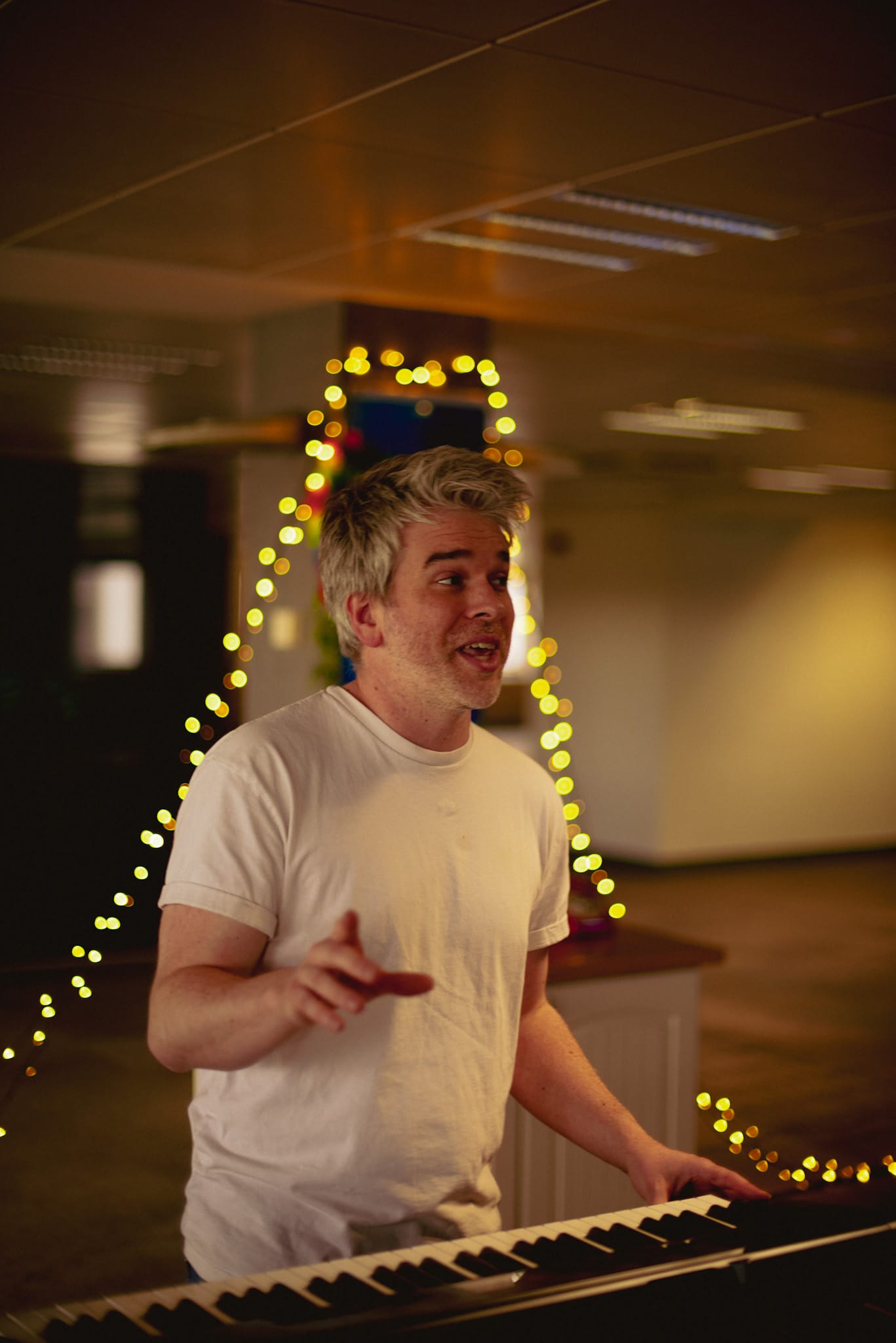 A performer in a white T-shirt plays keyboard in rehearsal, with string lights in the background.
