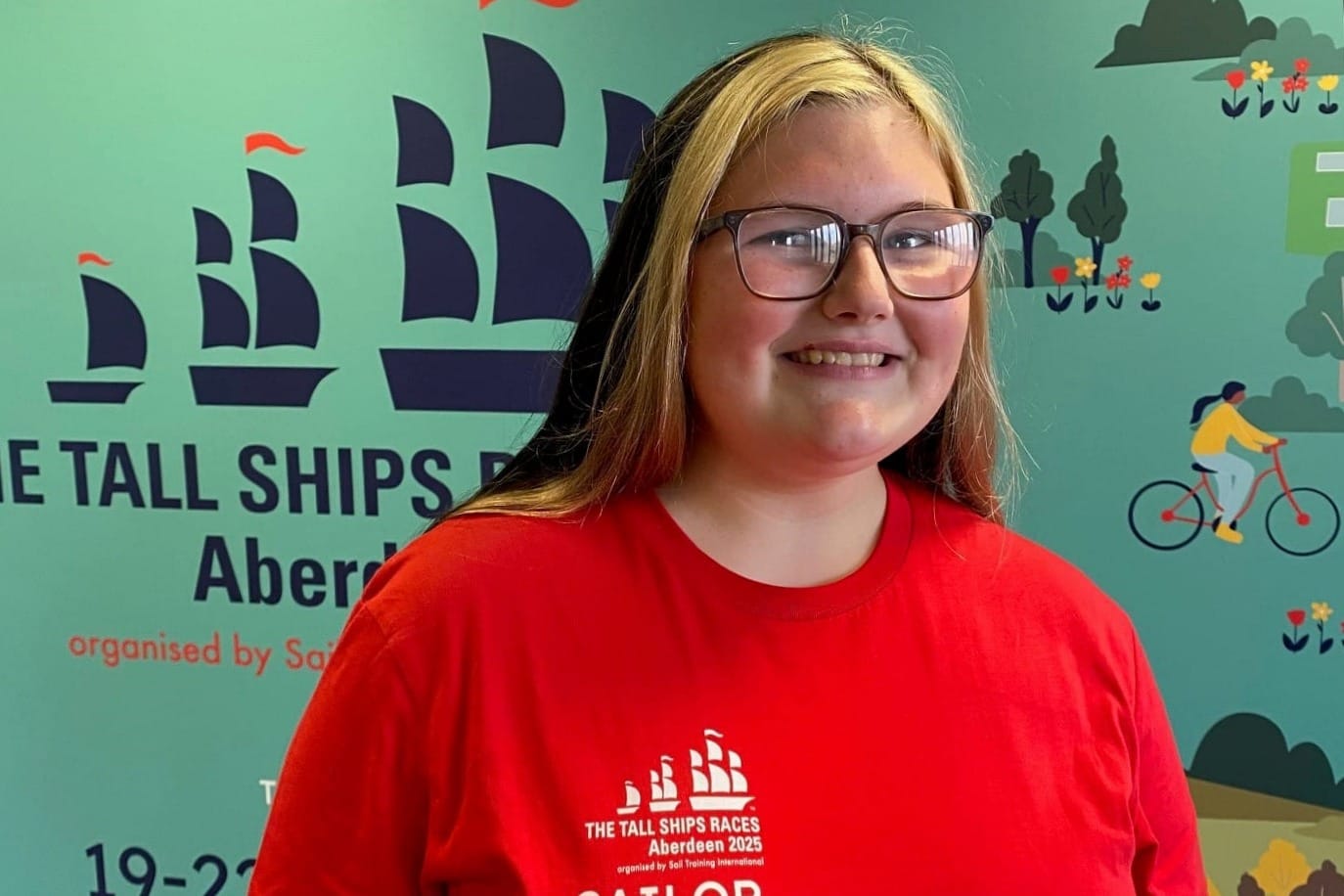 Maddie Sharpe smiles in front of a Tall Ships Races Aberdeen 2025 banner. She wears glasses and a red sailor t-shirt