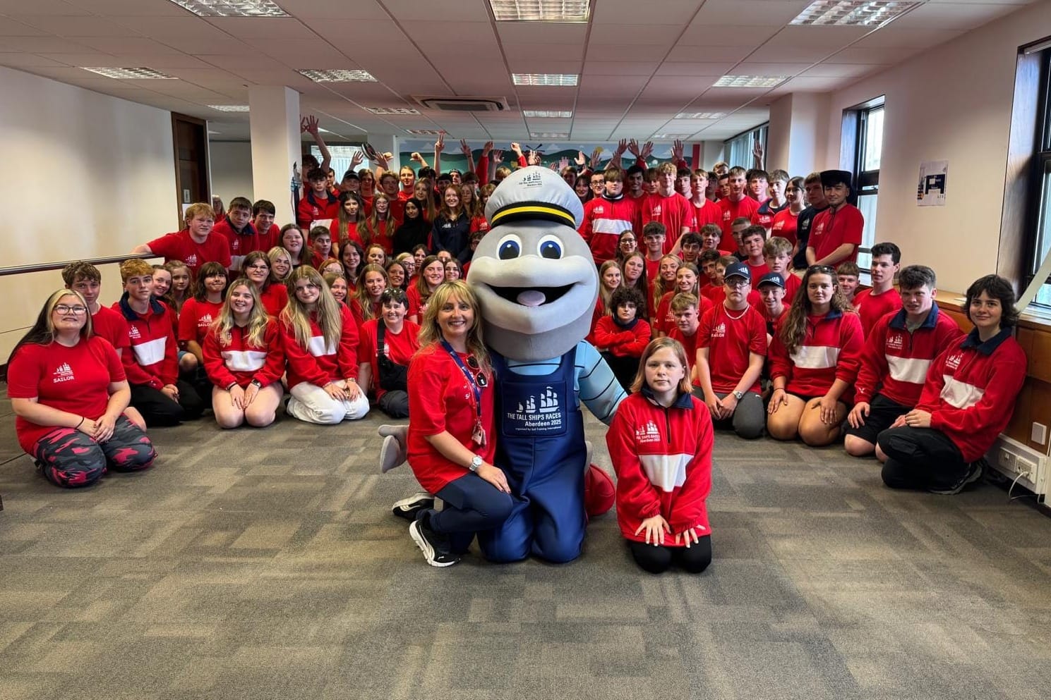 A large group of Tall Ships trainees in red uniforms pose indoors with a dolphin mascot and adult organisers