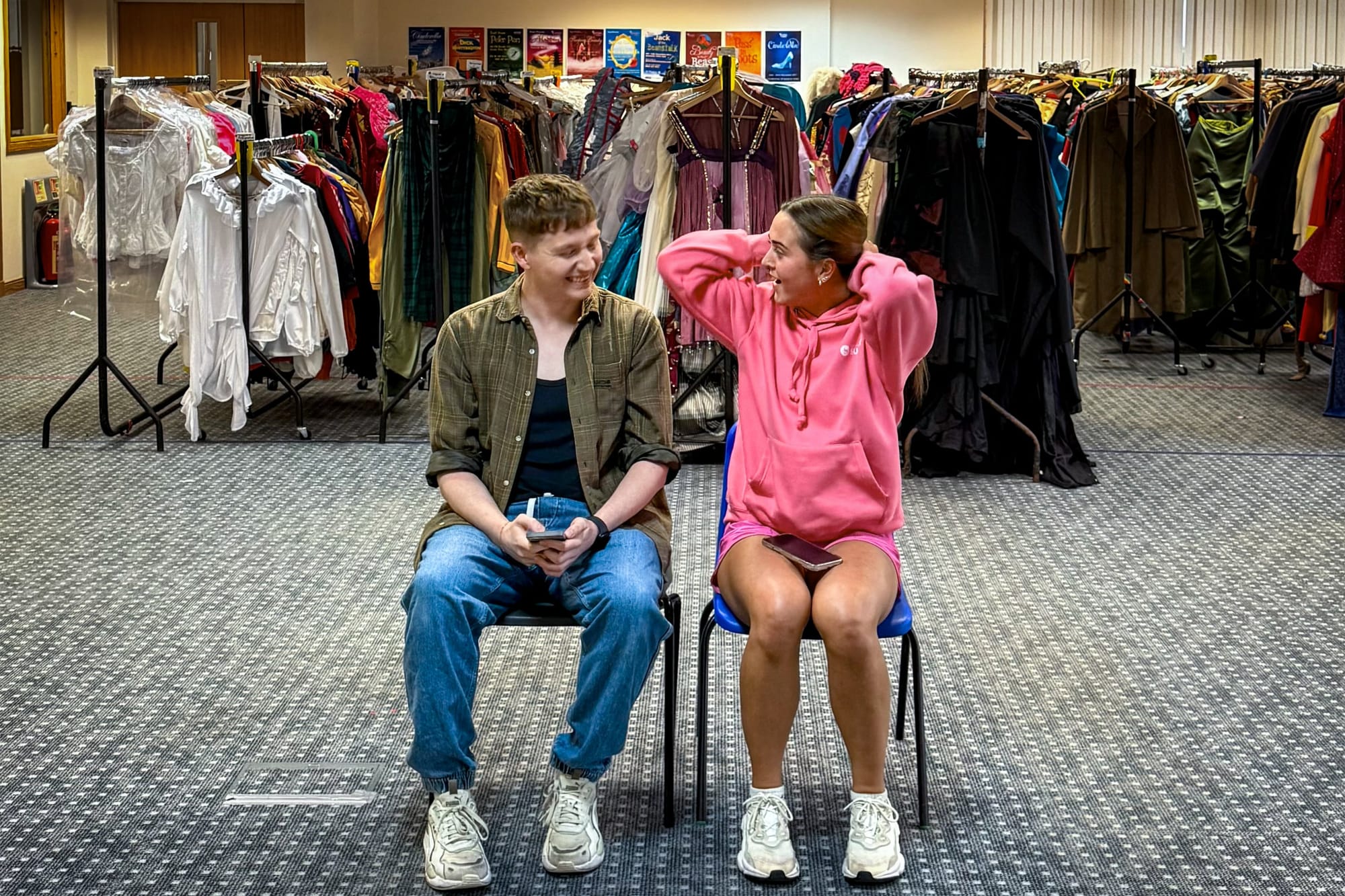 Two lead actors sit laughing in rehearsal with costume rails behind them.
