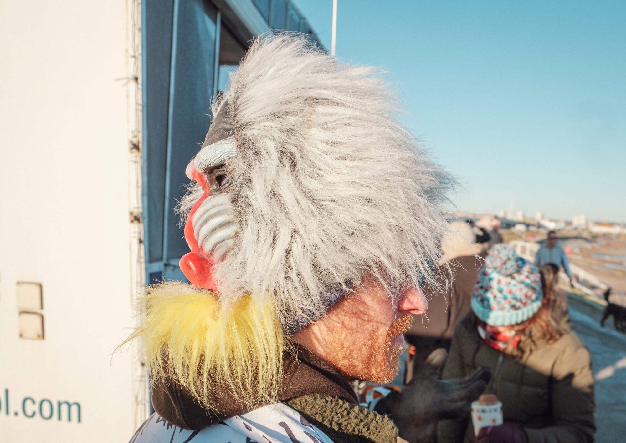 Side profile of man in furry baboon hat at a busy, cold beach.